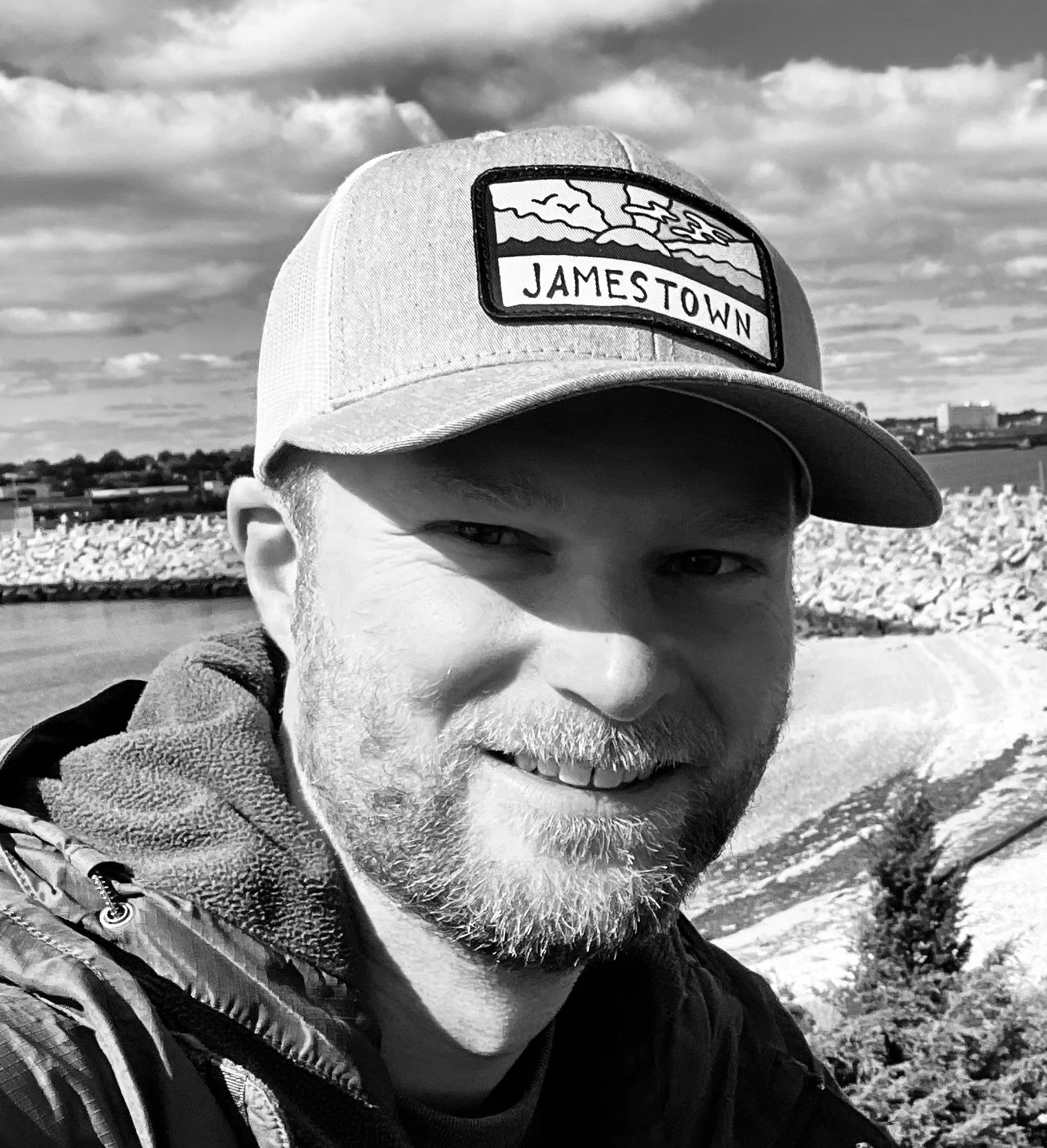 A man with a beard smiling outdoors, wearing a cap with a "JAMESTOWN" patch and a jacket, with a rocky embankment and a cloudy sky in the background.
