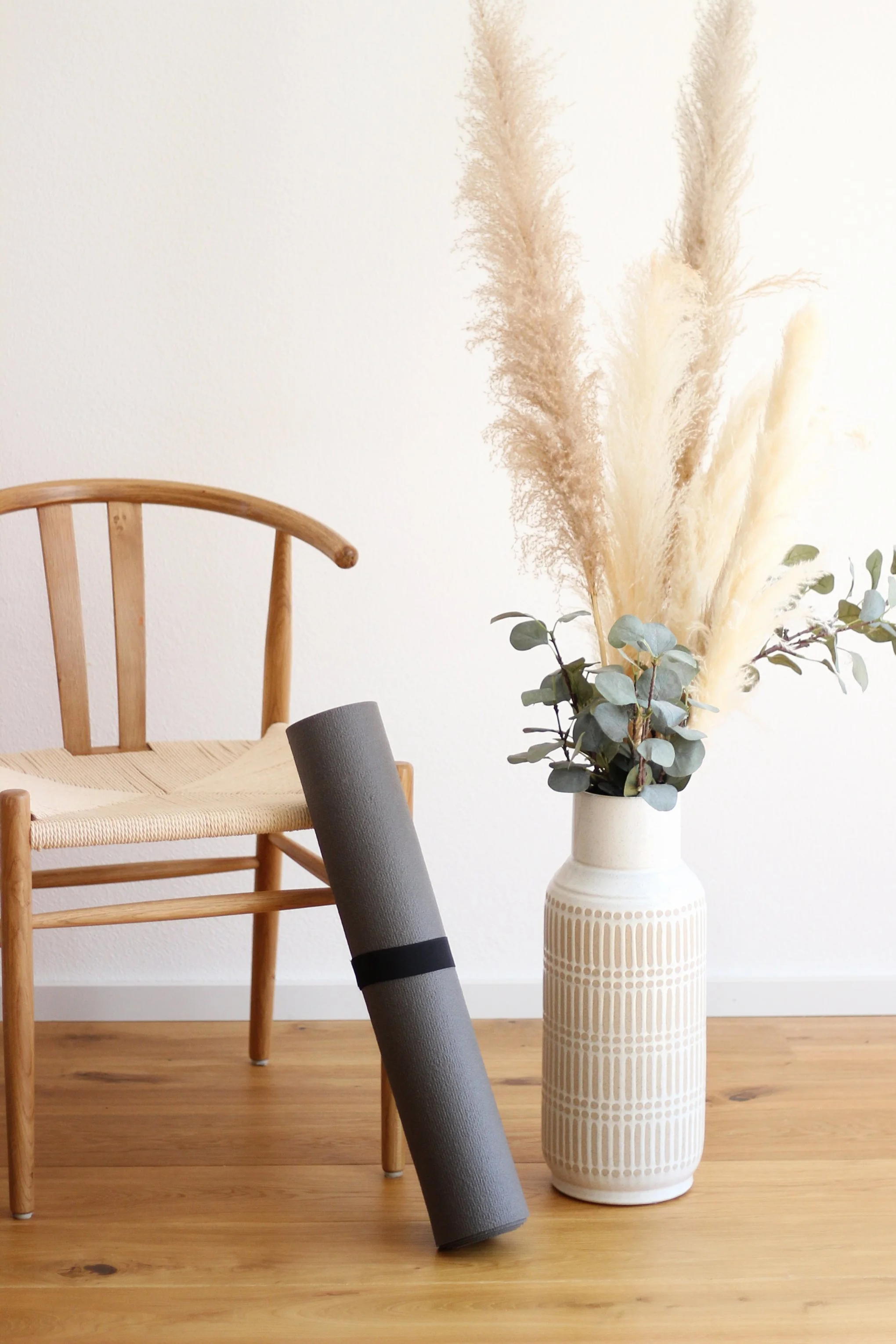 Ein Holzstuhl, eine Yogamatte, eine Vase mit getrockneten Pampas-Gräsern und Eukalyptus auf einem Holztisch vor einer weißen Wand.