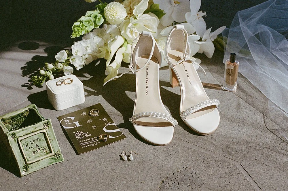 Bridal accessories including white high-heeled shoes with pearl embellishments, a pair of pearl earrings, gold rings, wedding veil, perfume bottle, wedding invitation, and floral arrangement on a gray surface.