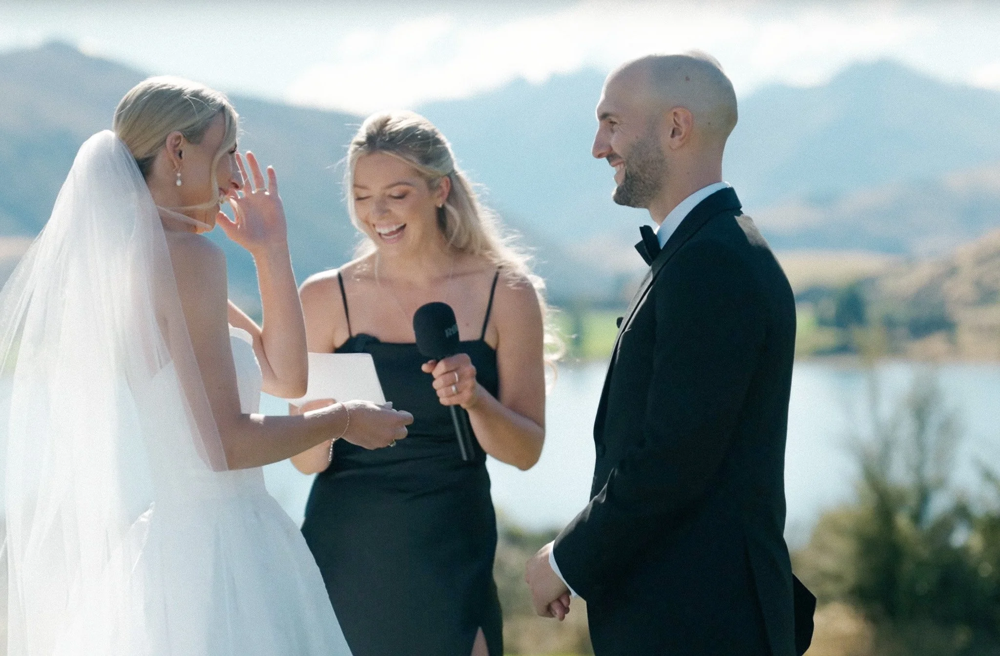 Bride reading vows during outdoor wedding ceremony as groom smiles and wedding officiant holds microphone.
