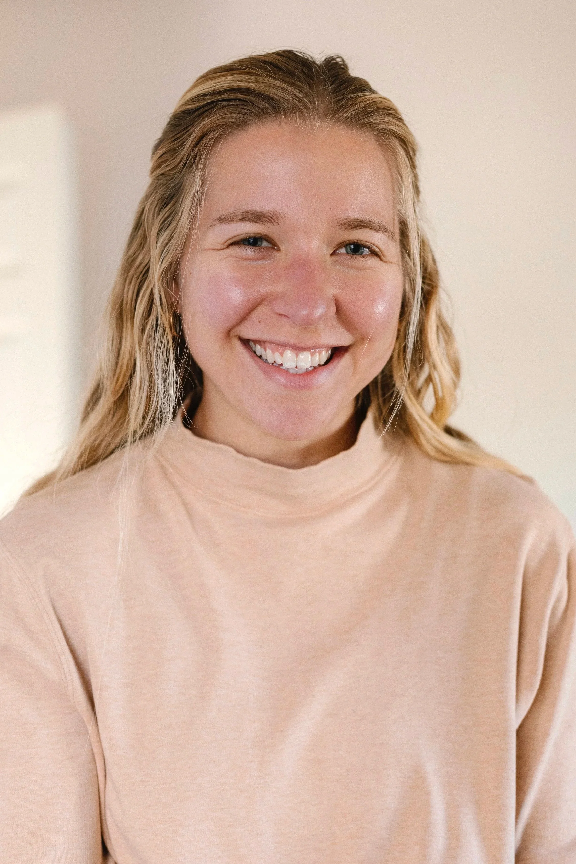 Close-up of a woman with blonde hair smiling at the camera, wearing a light beige sweater.