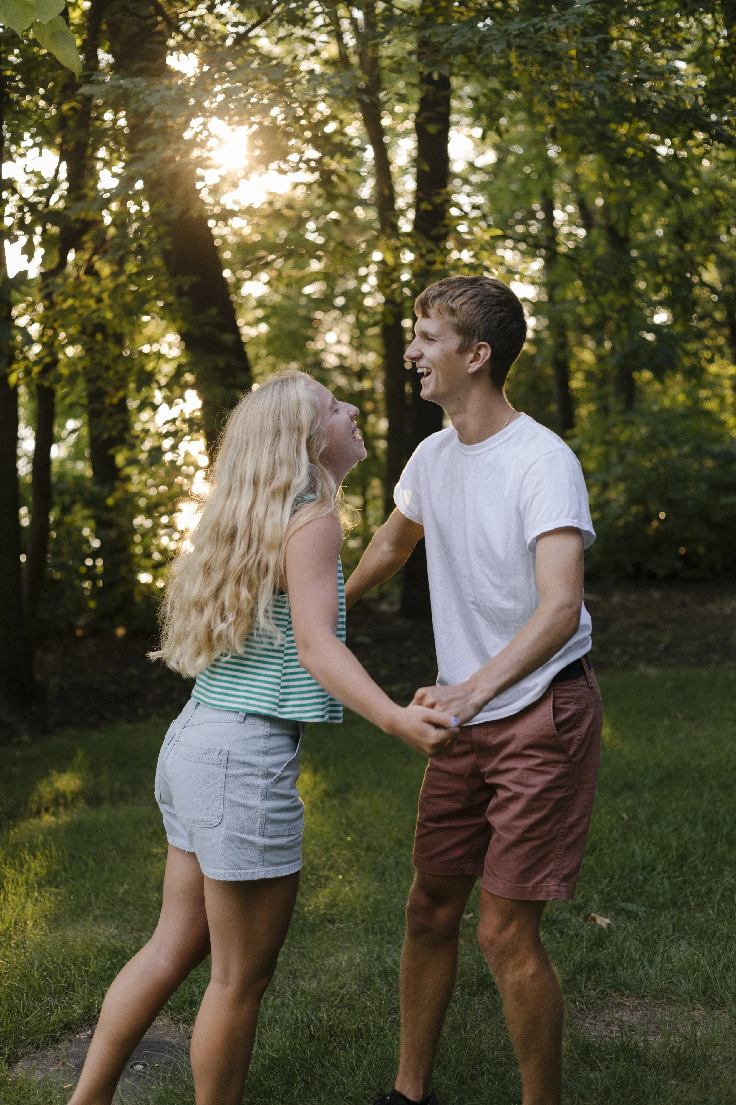A young couple engaging in a joyful moment outdoors in a green wooded area, holding hands and smiling at each other during sunset.