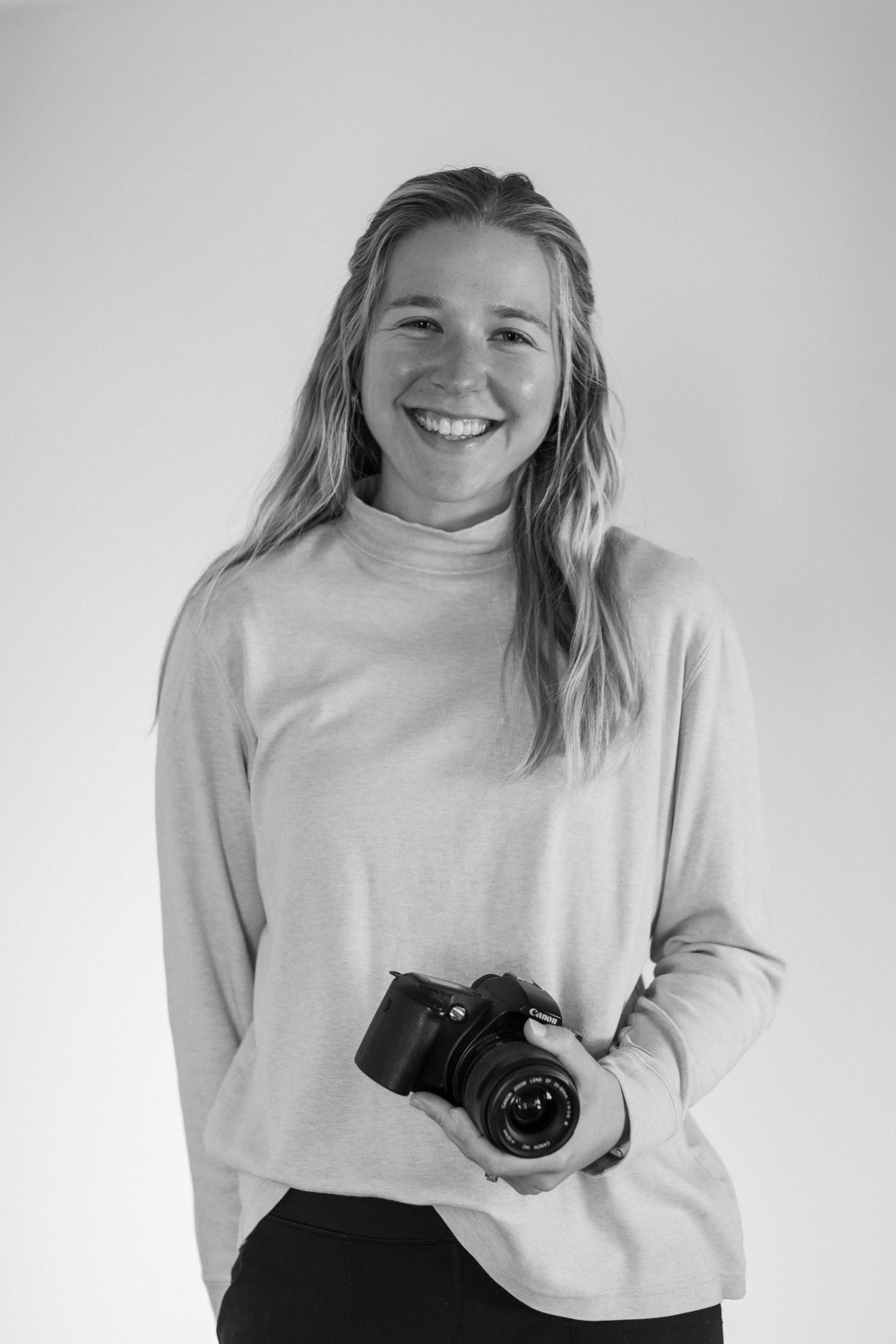 A smiling woman with blonde hair holding a camera in her right hand, wearing a light-colored long-sleeve shirt, against a plain background.