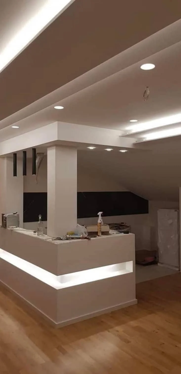 Intérieur d'une pièce en cours de rénovation avec un plafond suspendu, des spots encastrés, un comptoir de cuisine blanc avec des outils et des matériaux, et un sol en parquet.