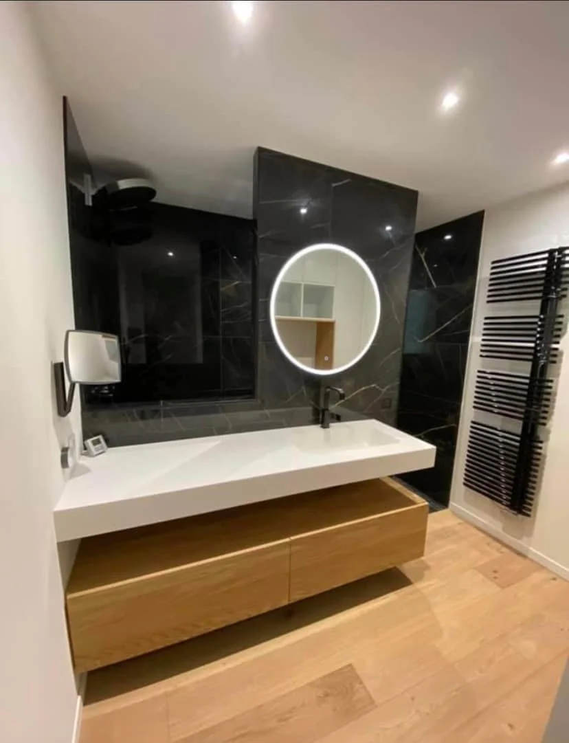 Une salle de bain moderne avec un lavabo à mi-hauteur sur une caisse en bois, un miroir rond lumineux, des murs en carrelage noir, un radiateur noir vertical, et un sol en bois clair.