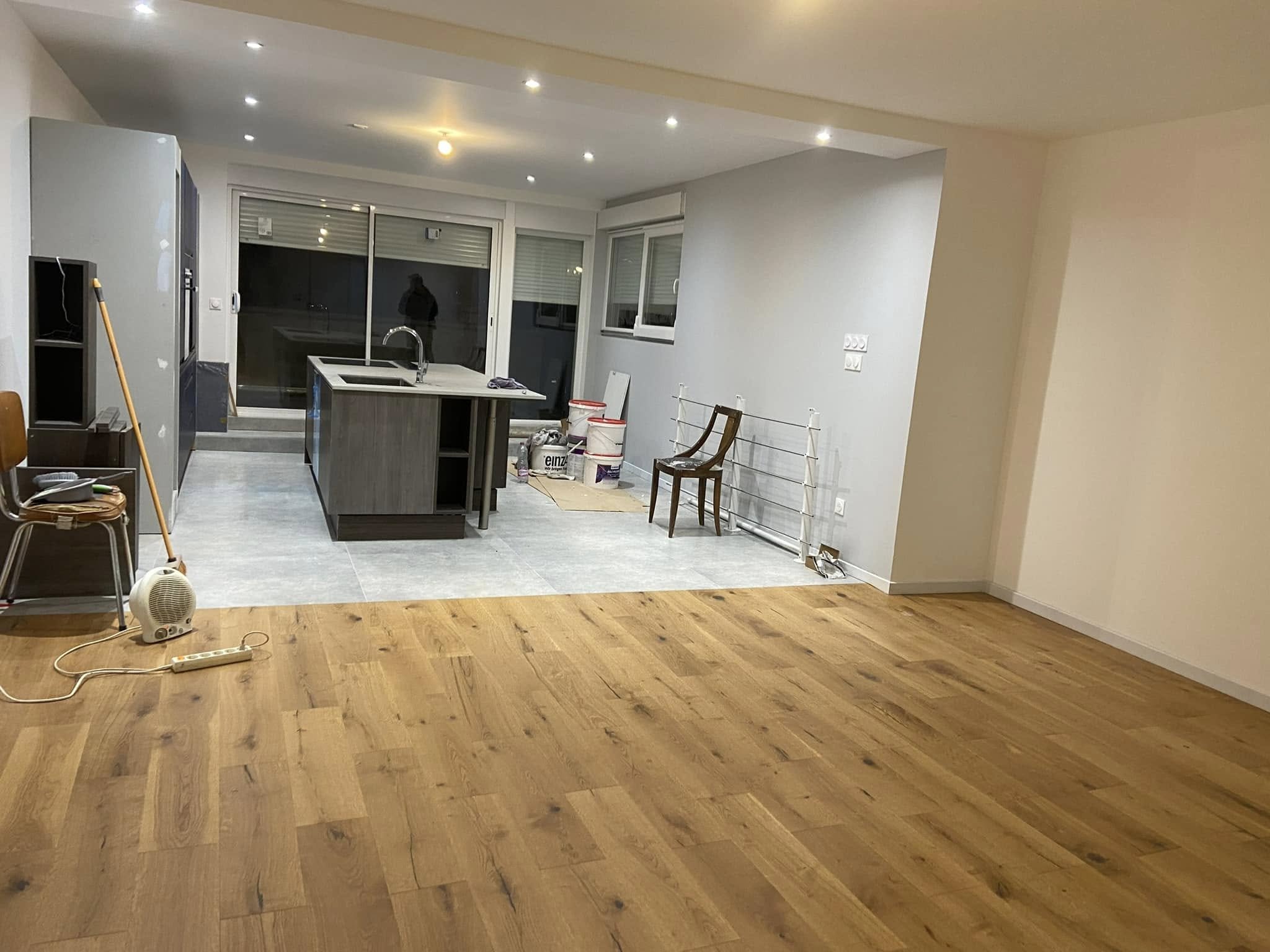 Une pièce en cours de rénovation avec un sol en bois, une cuisine moderne avec un îlot, des chaises en bois, des buckets et des outils de rénovation contre le mur.