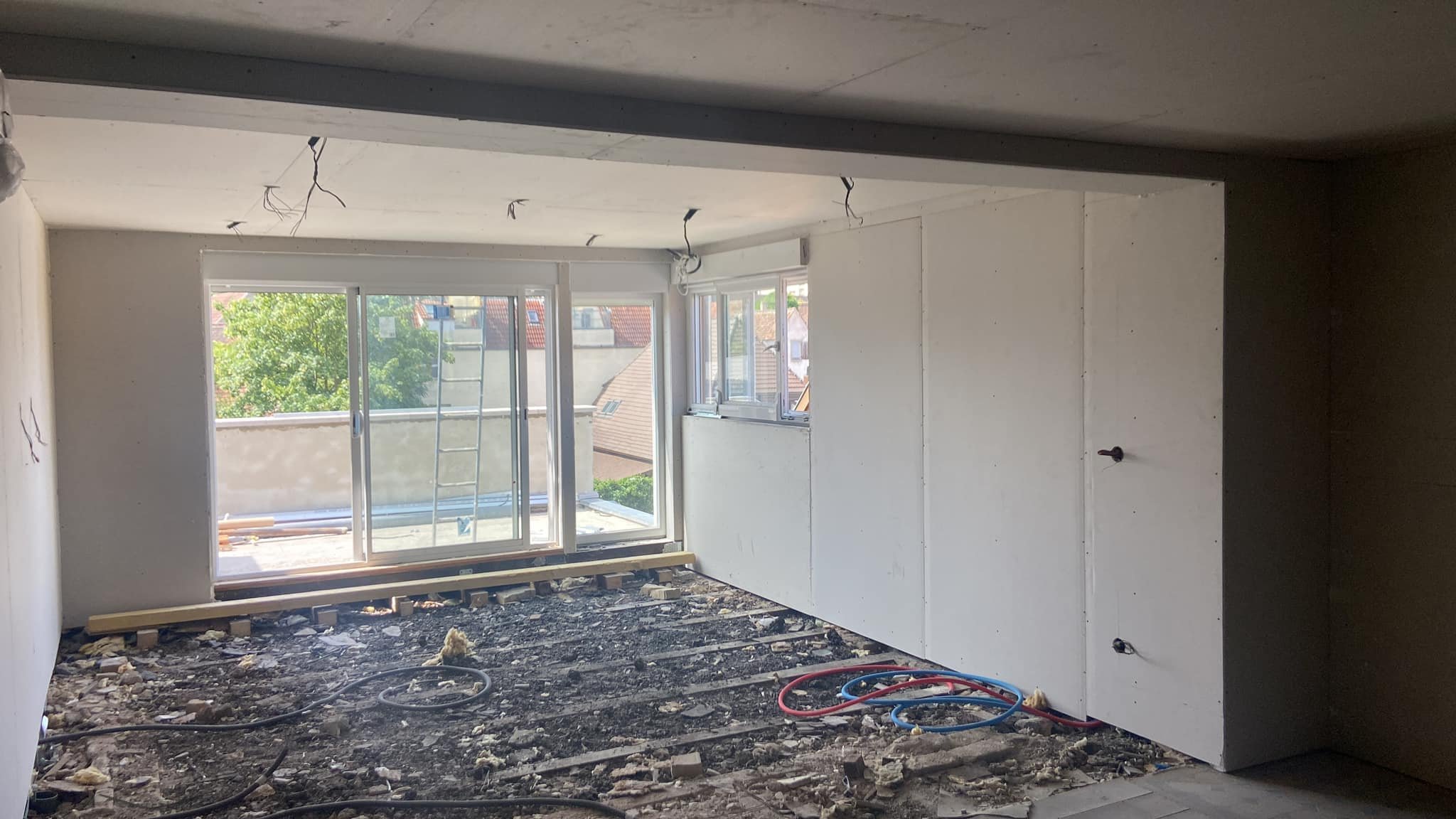 Intérieur d'une pièce en cours de rénovation, avec murs en plâtre, sol en travaux, et fenêtre donnant sur un balcon avec des toits et arbres en arrière-plan.