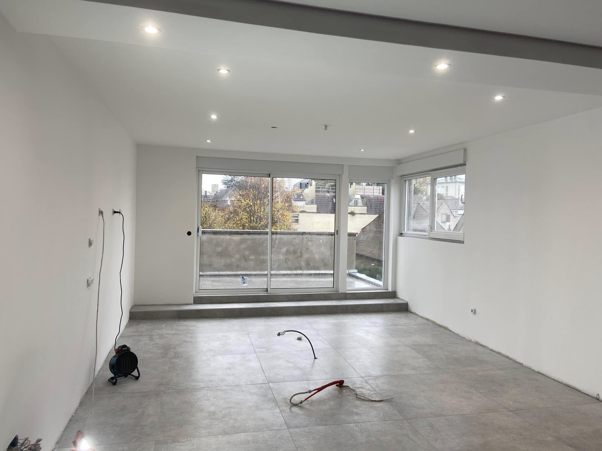 Salle de séjour en cours de rénovation avec murs blancs, sol en carrelage, vues sur un balcon et des fenêtres offrant une lumière naturelle. Éléments électriques en cours d'installation.