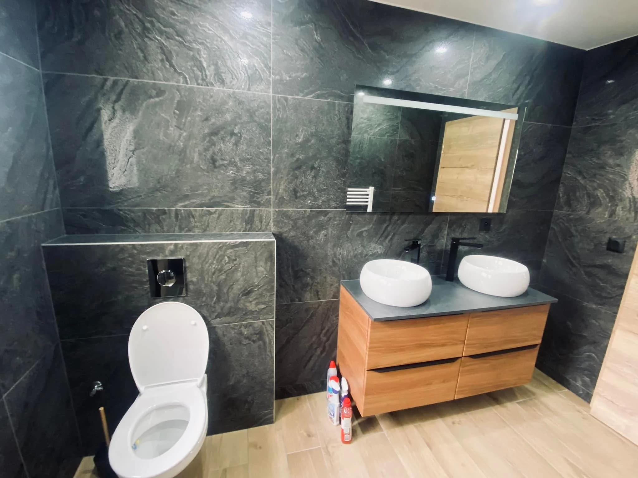 Salle de bain moderne avec deux lavabos ovales en céramique blanche sur un meuble en bois, grand miroir rectangulaire, toilettes, murs en pierre grise, sol en carrelage beige, produits de nettoyage sous le meuble.