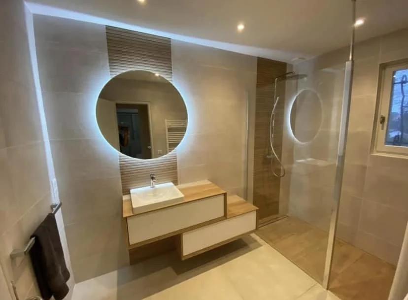 Salle de bain moderne avec un miroir rond éclairé, évier sur un meuble en bois, douche séparée avec porte en verre, fenêtre, et un éclairage intégré au plafond.