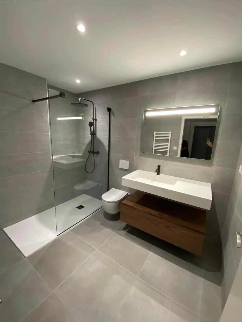 Salle de bain moderne avec douche à l'italienne, lavabo à surface plane avec meuble en bois, miroir lumineux et toilette suspendue, murs et sol carrelés gris.