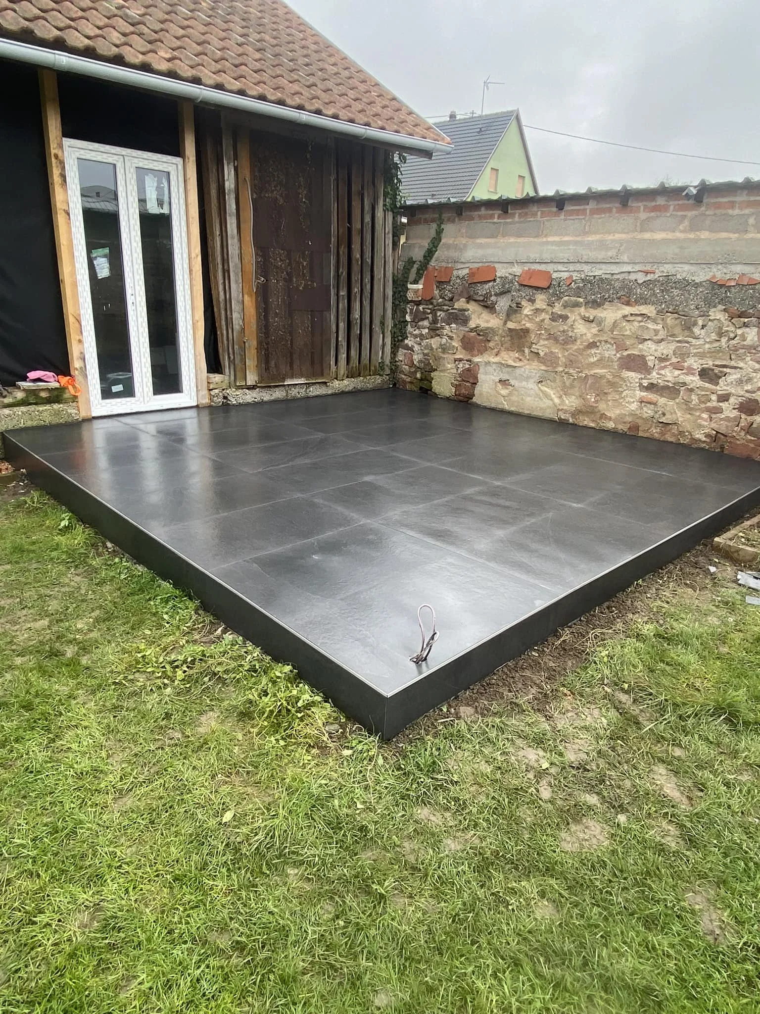 Terrasse en pierre noire fraîchement rénovée à côté d'une vieille clôture en pierre et bois, avec une maison en arrière-plan.