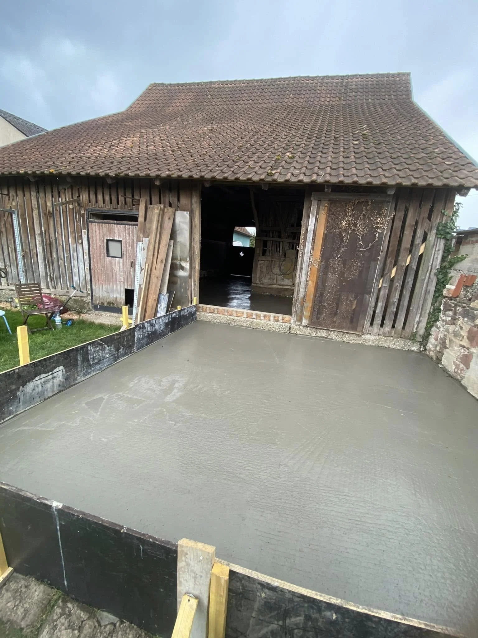 Une chape de béton fraîchement coulée devant une vieille structure en bois, probablement un abri ou un garage, avec un ciel partiellement nuageux.