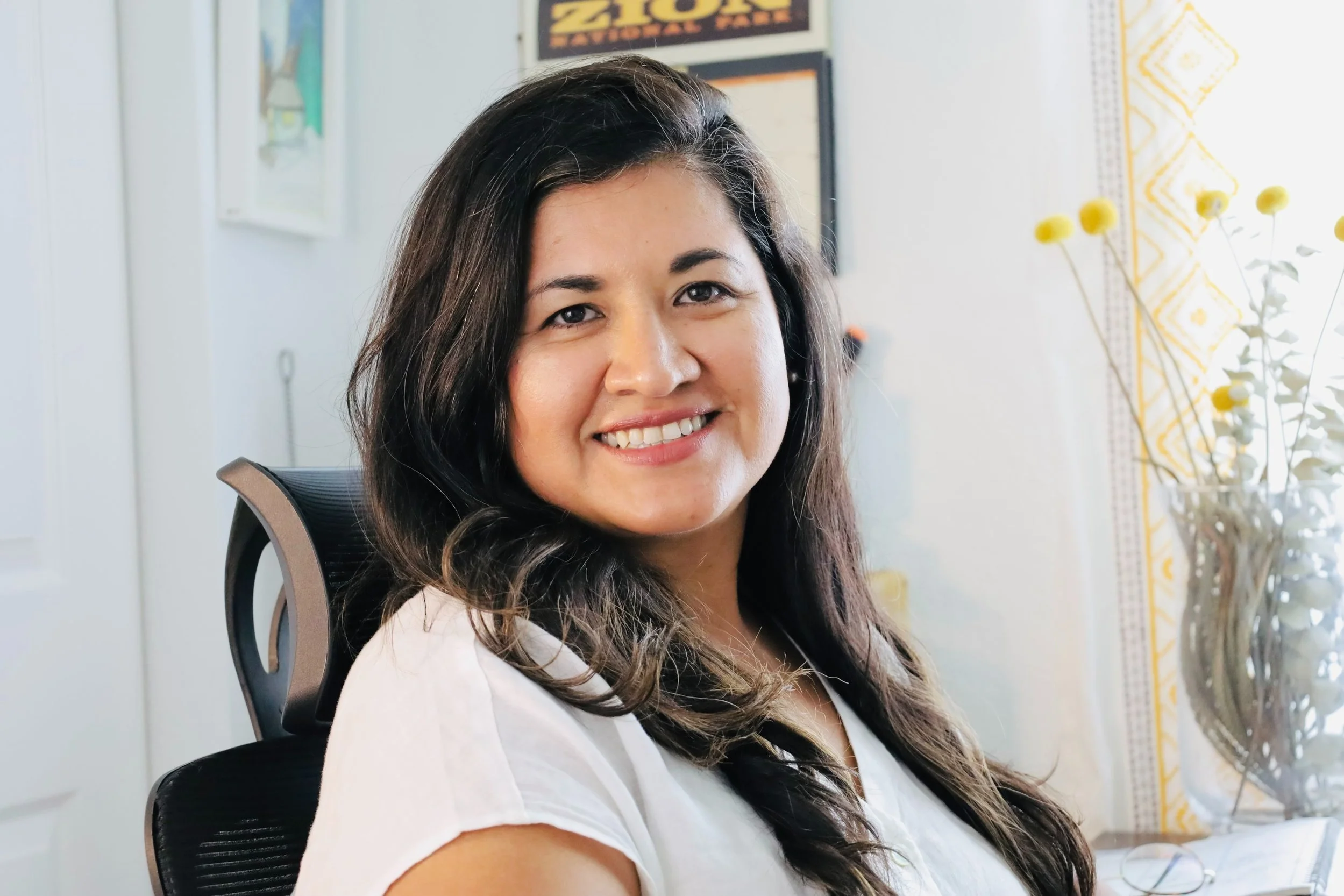 A woman with long dark hair smiling, sitting in a black office chair in a well-lit room with white walls, yellow decorative curtains, a vase with white and yellow flowers, and framed posters on the wall.