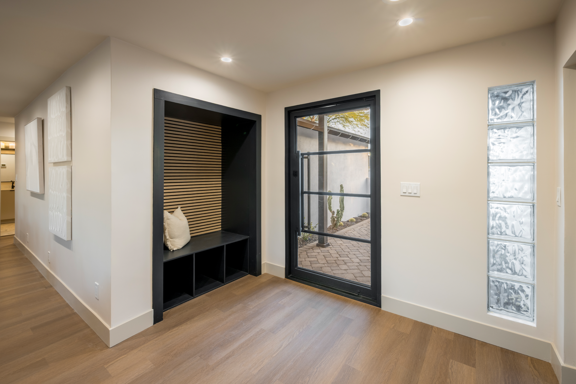 Interior view of a modern room with a glass door leading outside, a built-in black nook with a wood slat back, and glass block wall on the right, with white walls and wooden flooring.