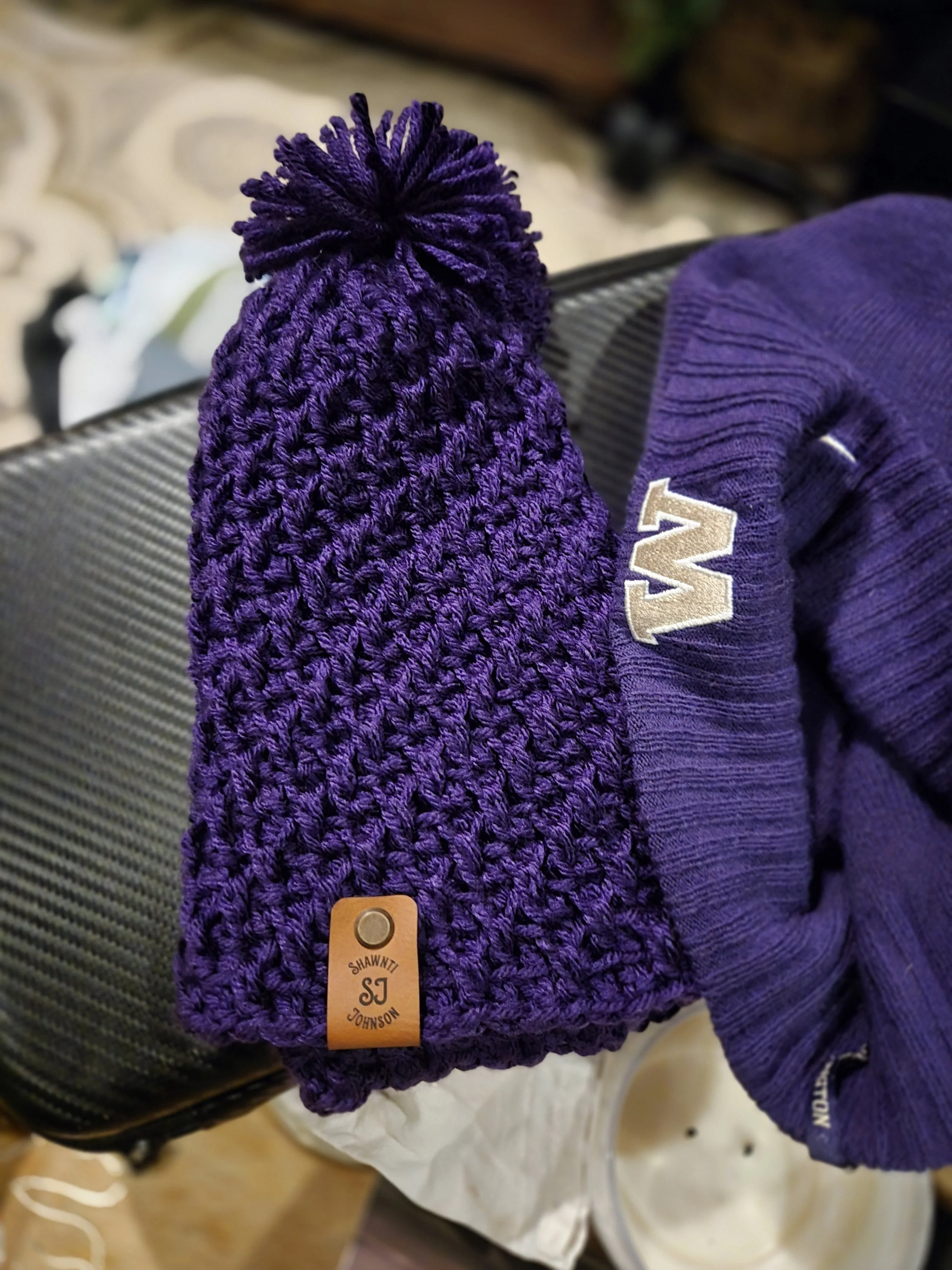 Purple knit beanie with pom-pom and leather label next to a purple knitted hat with a letter 'W' logo.