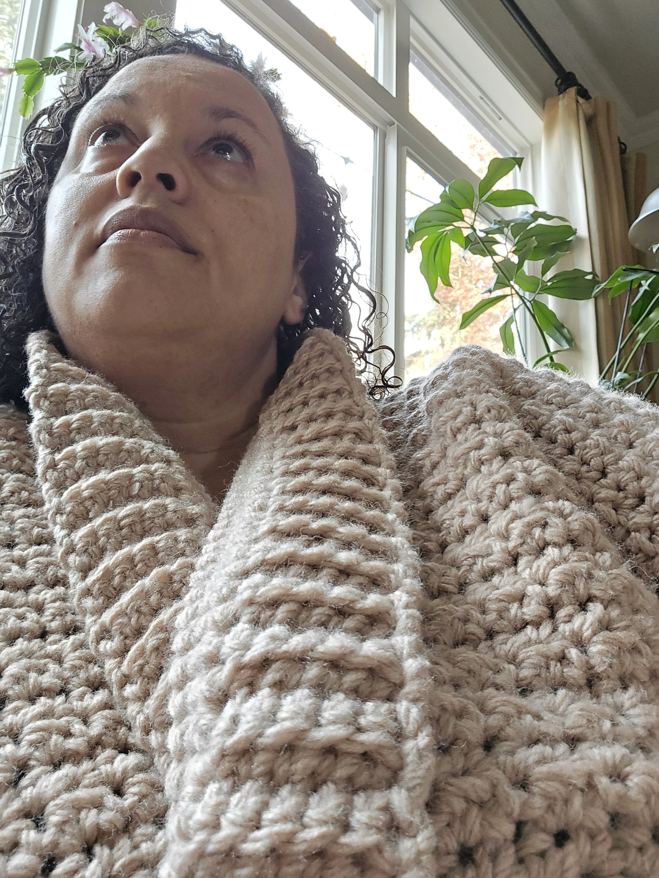 Woman wearing a textured beige sweater sits near a window with green plants in the background.