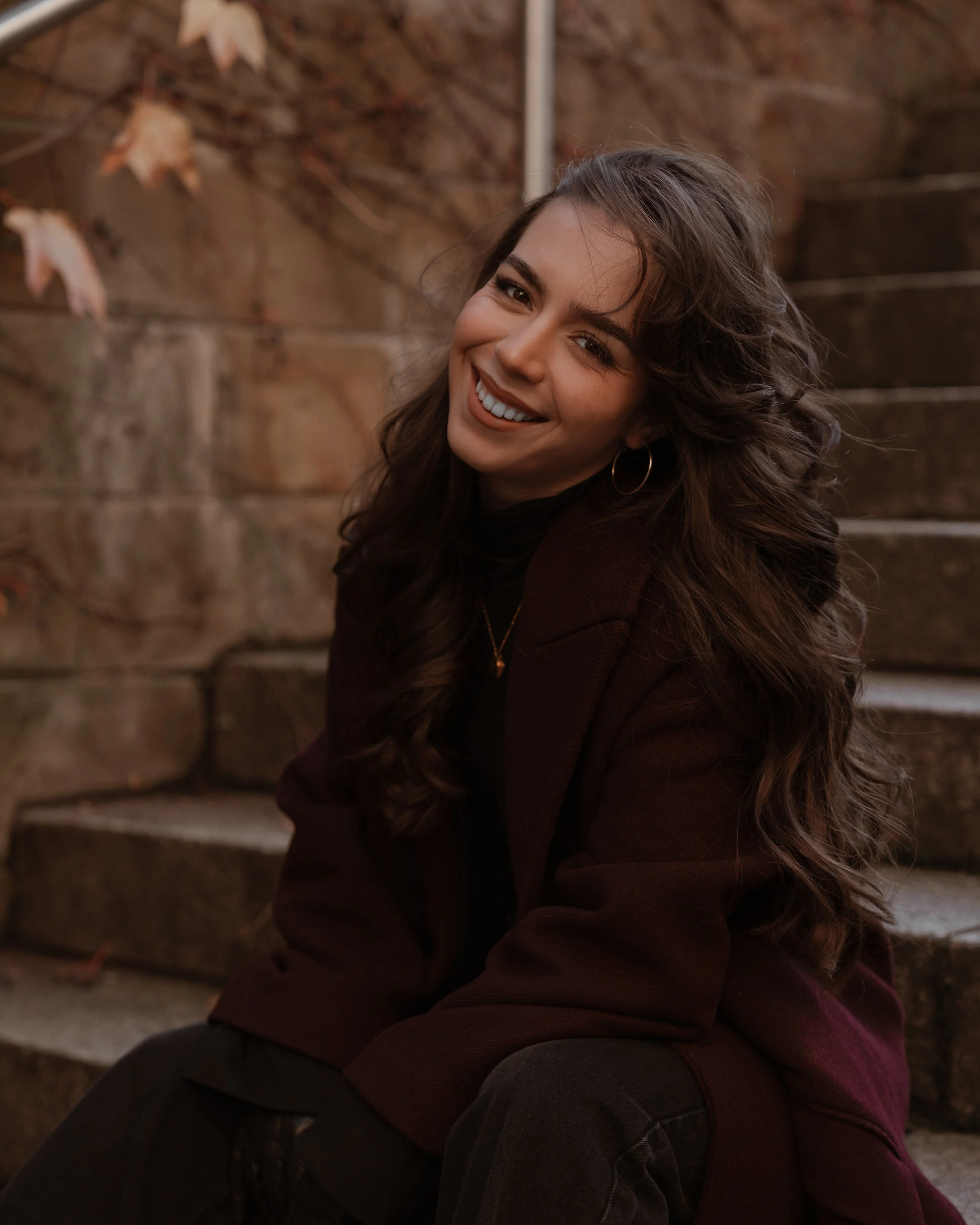 Junge Frau mit langen, lockigen braunen Haaren lächelnd, trägt eine schwarze Top und braune Jacke, sitzt auf Steinstufen, im Hintergrund eine Wand mit Herbstlaub, lacht freundlich.