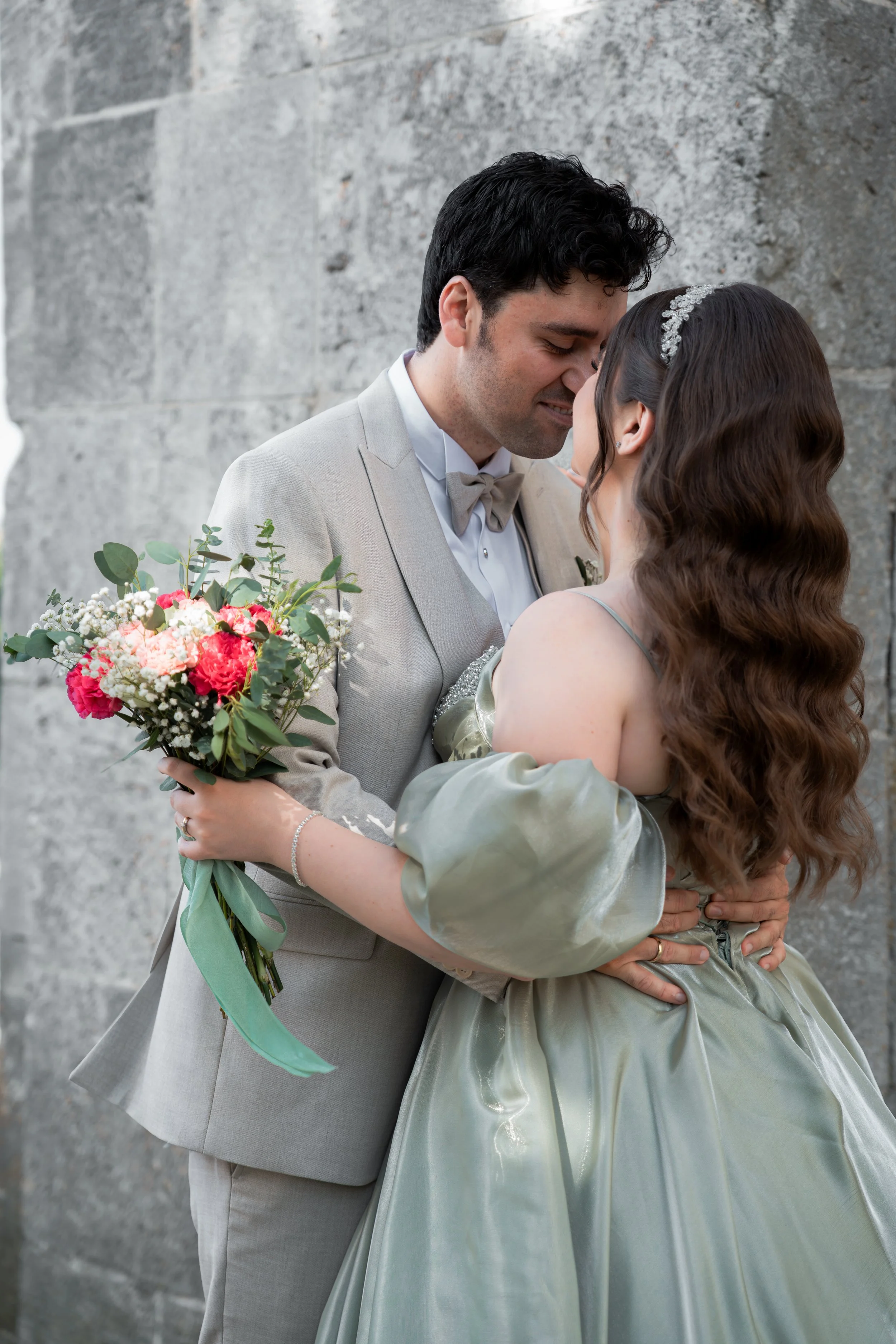 Ein Paar bei einer Hochzeitszeremonie, das sich liebevoll umarmt, vor einer Steinmauer, mit Blumenstrauß in der Hand.