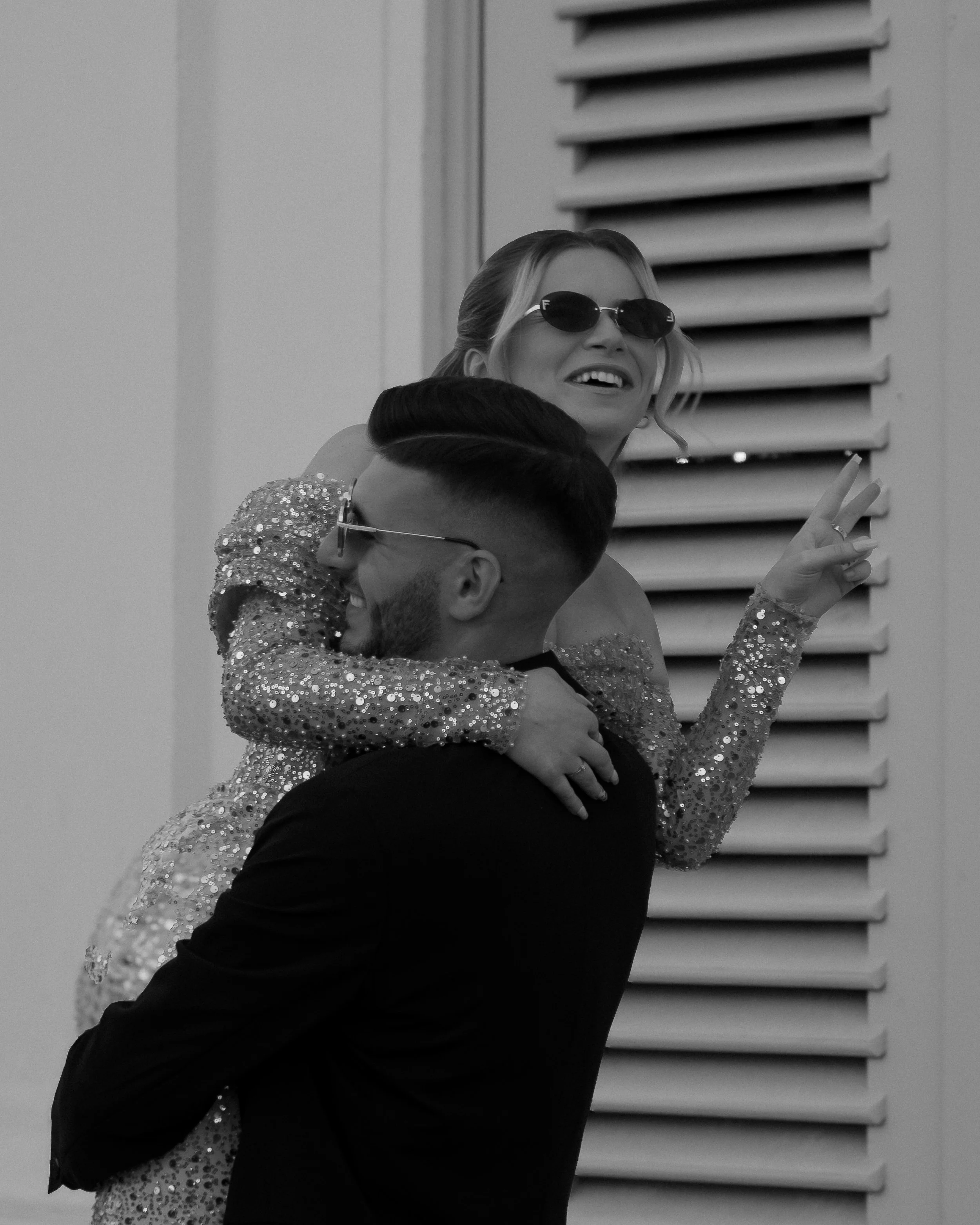 Ein Mann hebt eine Frau hoch; beide tragen Sonnenbrillen. Die Frau zeigt das Peace-Zeichen und sie lächeln. Sie trägt ein glitzerndes Kleid, und der Mann ist in dunkler Kleidung gekleidet.