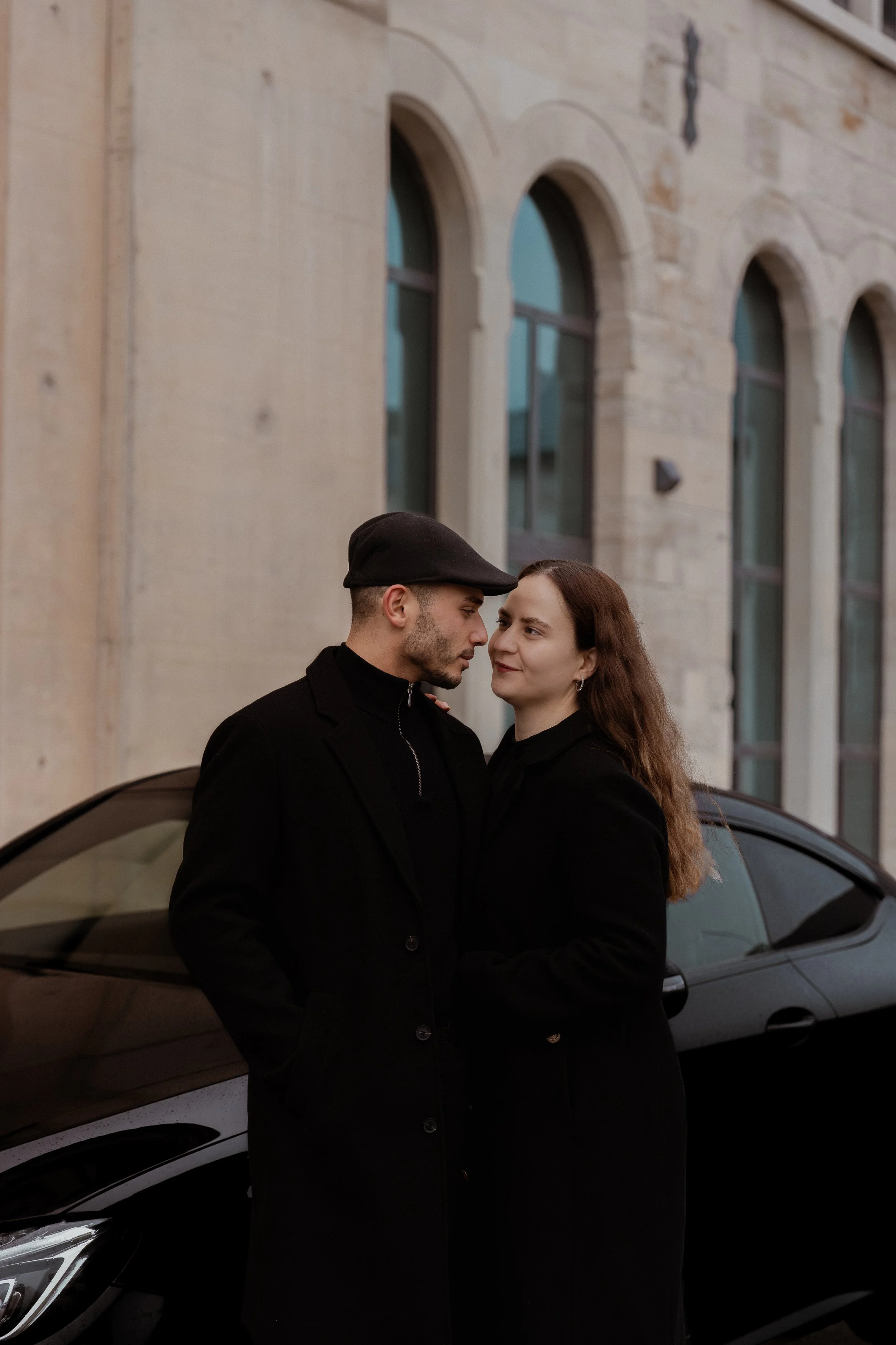 Ein Mann und eine Frau stehen eng beieinander vor einem schwarzen Auto und schauen sich liebevoll an, im Hintergrund ein altes Steingebäude mit großen Fenstern.