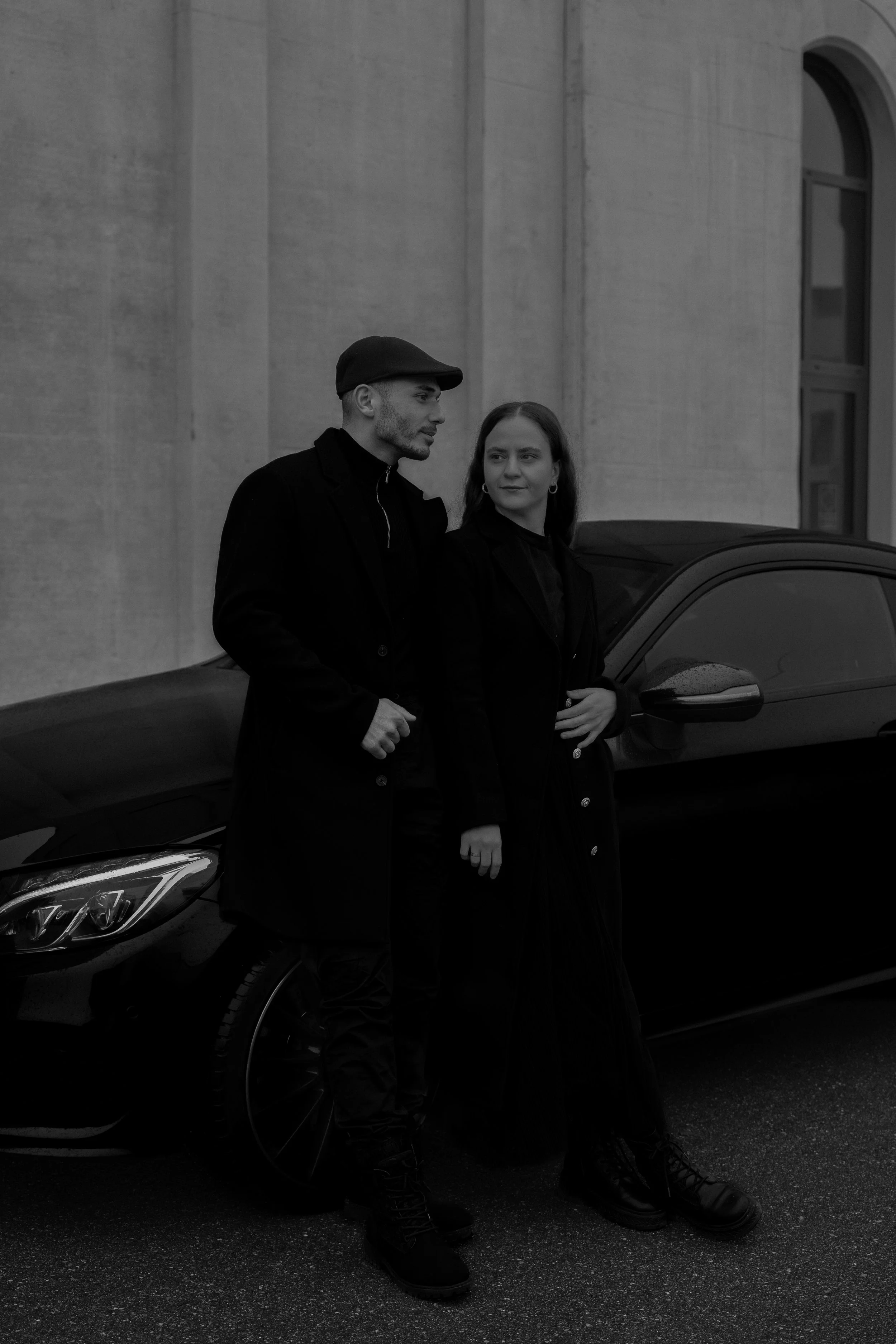 Ein Mann und eine Frau in schwarzer Kleidung stehen nebeneinander vor einem Auto, das an einer Betonwand geparkt ist.