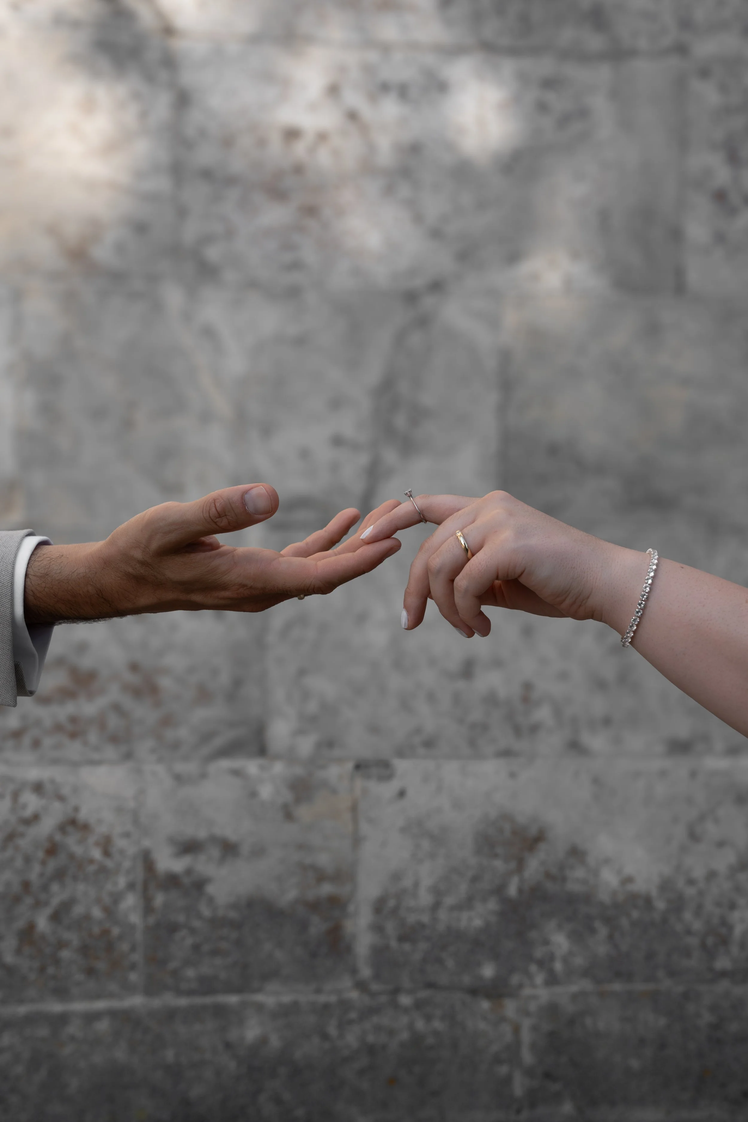 Ein Mann übergibt einer Frau einen Verlobungsring, beide Hände sind sichtbar, die Frau trägt eine Diamantenarmband. Der Hintergrund ist eine Steinmauer.
