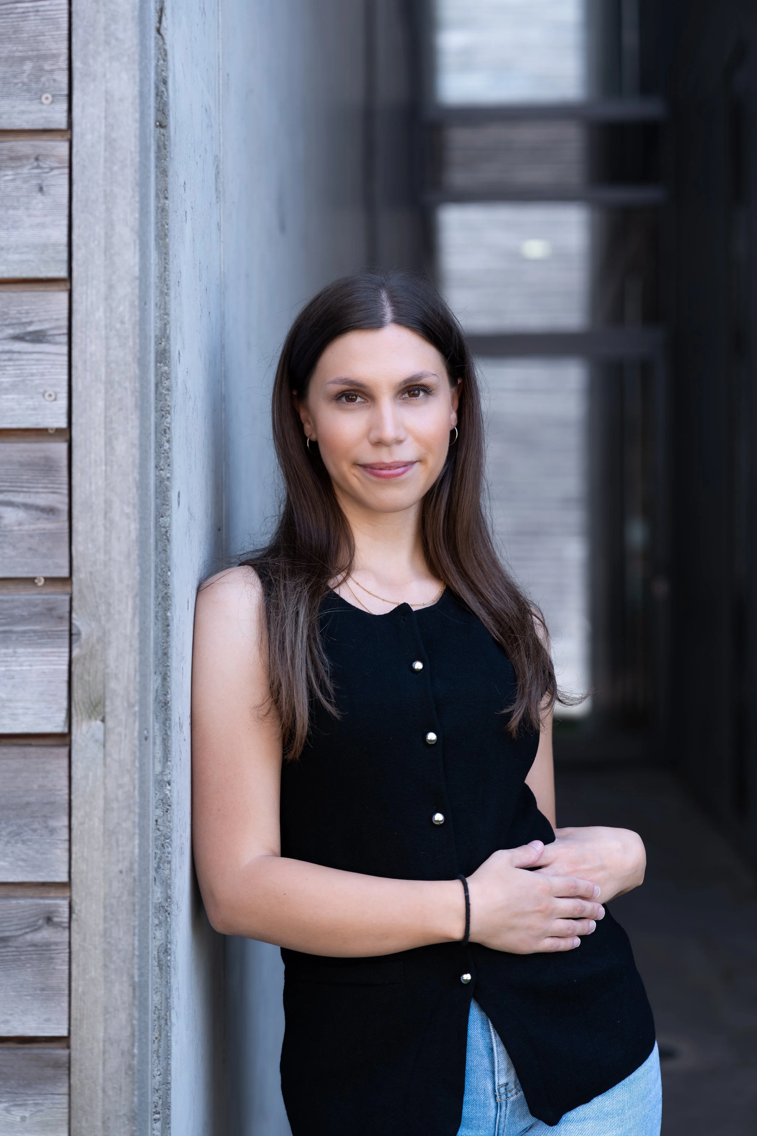 Junge Frau mit braunen Haaren und Schmuck lehnt an einer Wand, trägt ein schwarzes Top mit Perlenknöpfen und jeans, im Hintergrund eine moderne, dunkle Architektur.