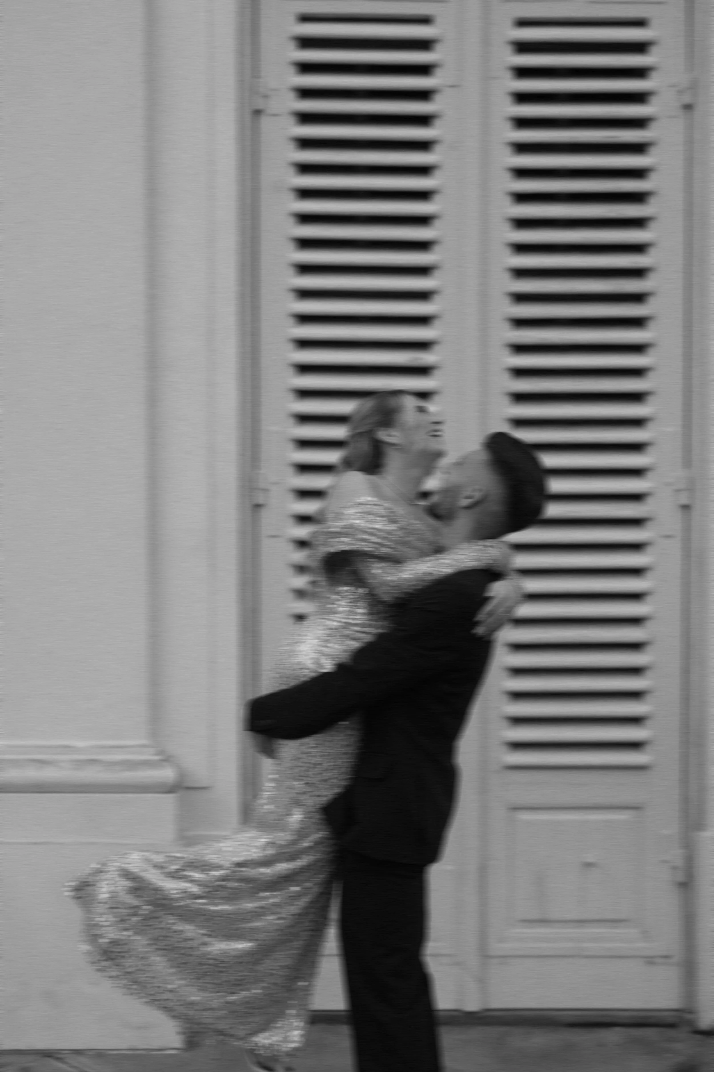 Ein Paar tanzt eng umschlungen vor einer weißen Tür mit Lamellen, beide tragen elegante Kleidung, sie trägt ein glitzerndes Kleid, er einen schwarzen Anzug.