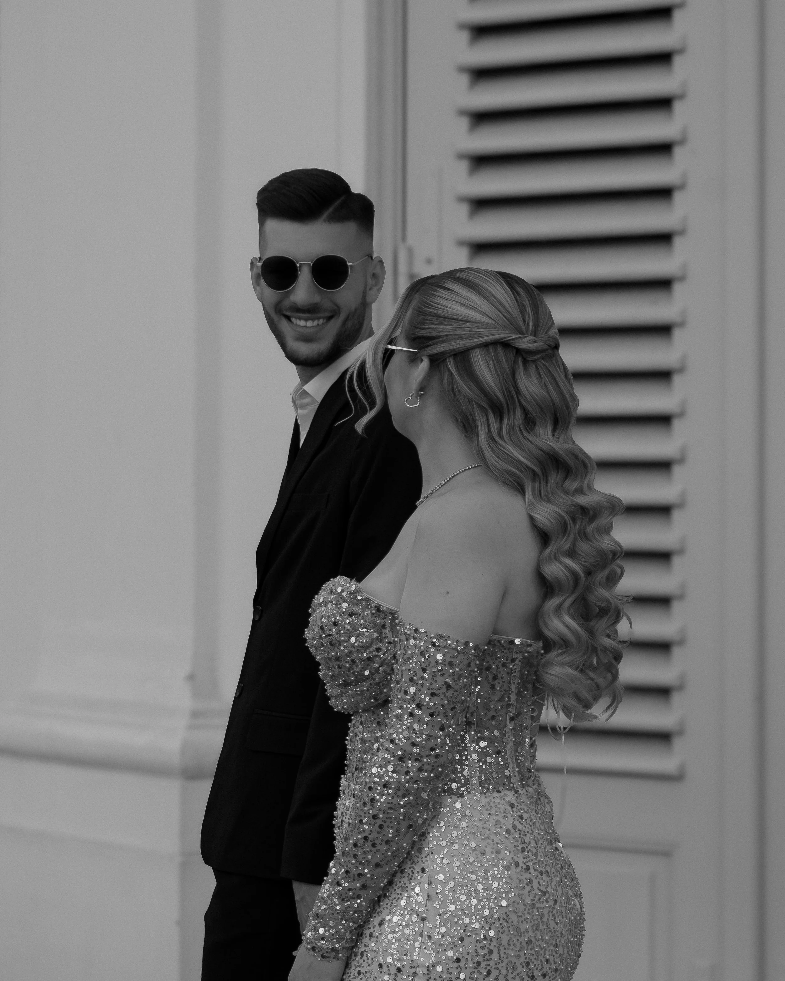 Ein Mann in Anzug und Sonnenbrille lächelt, während er neben einer Frau im glitzernden Kleid mit lockigem Haar steht. Beide stehen vor einem Gebäude mit Fensterläden.