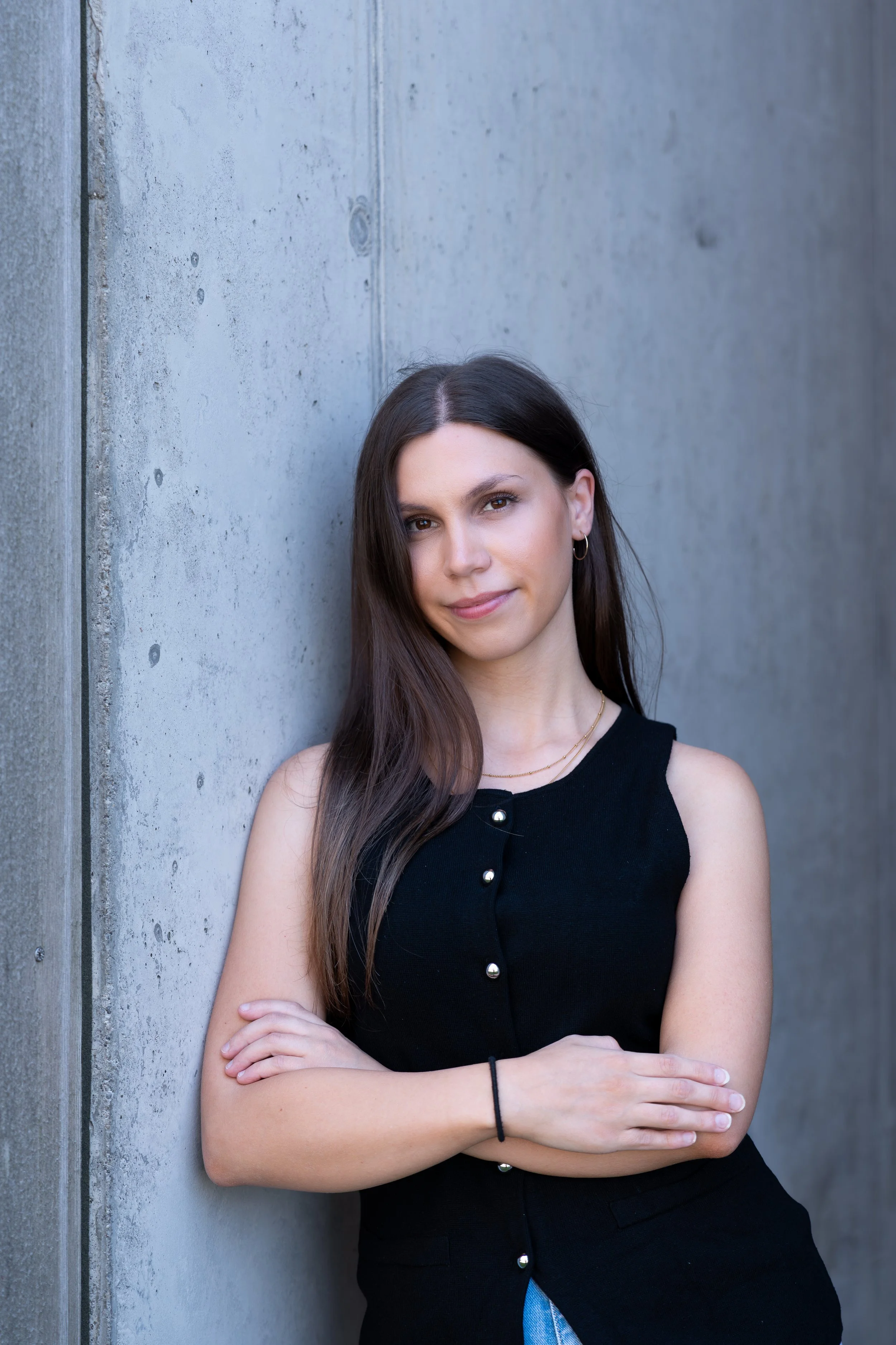 Junge Frau mit dunklen Haaren, schwarze Weste, lehnt an einer Betonwand und schaut in die Kamera.
