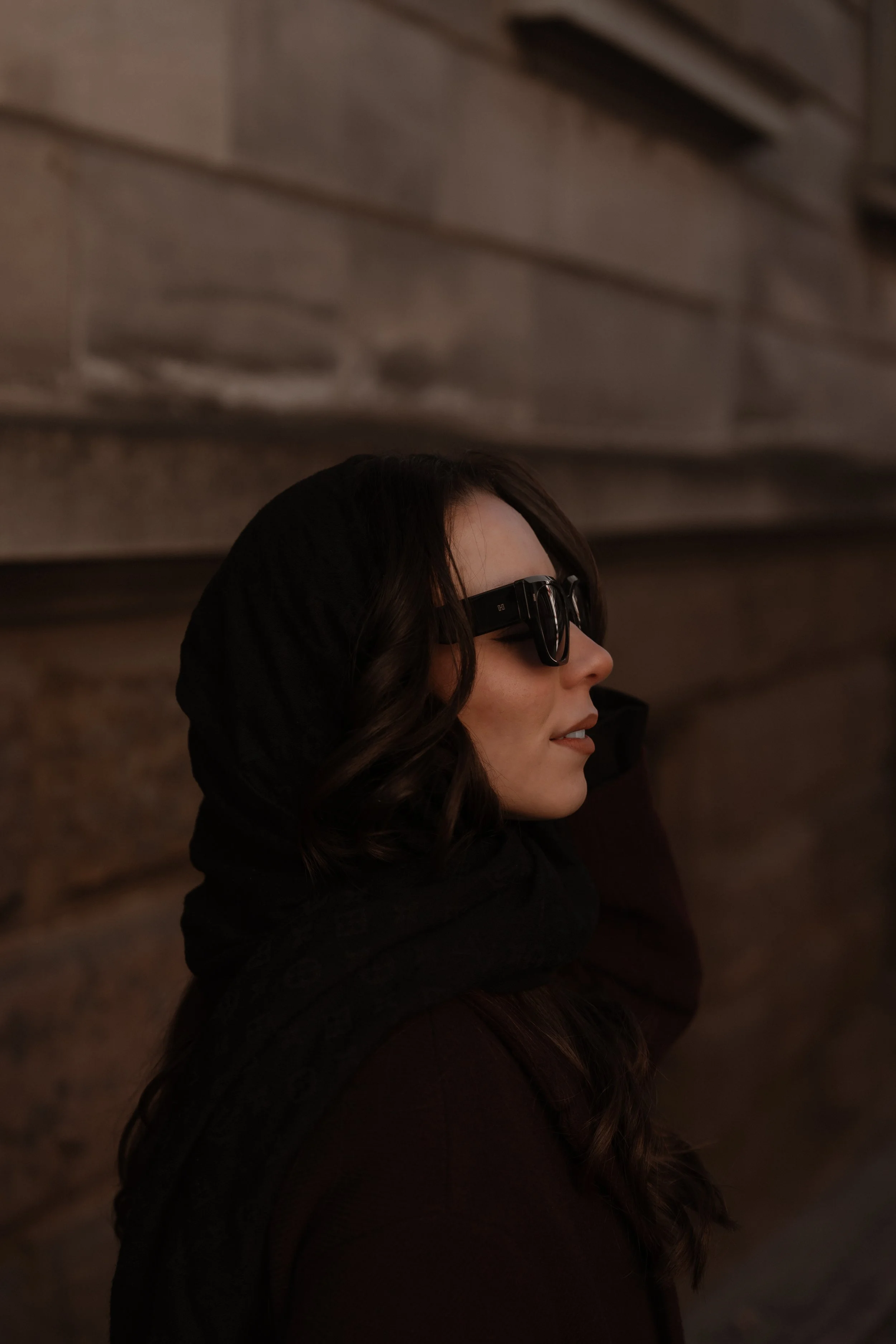 Frau mit schwarzen Haaren, Sonnenbrille und schwarzem Hoodie steht an einer Steinmauer.