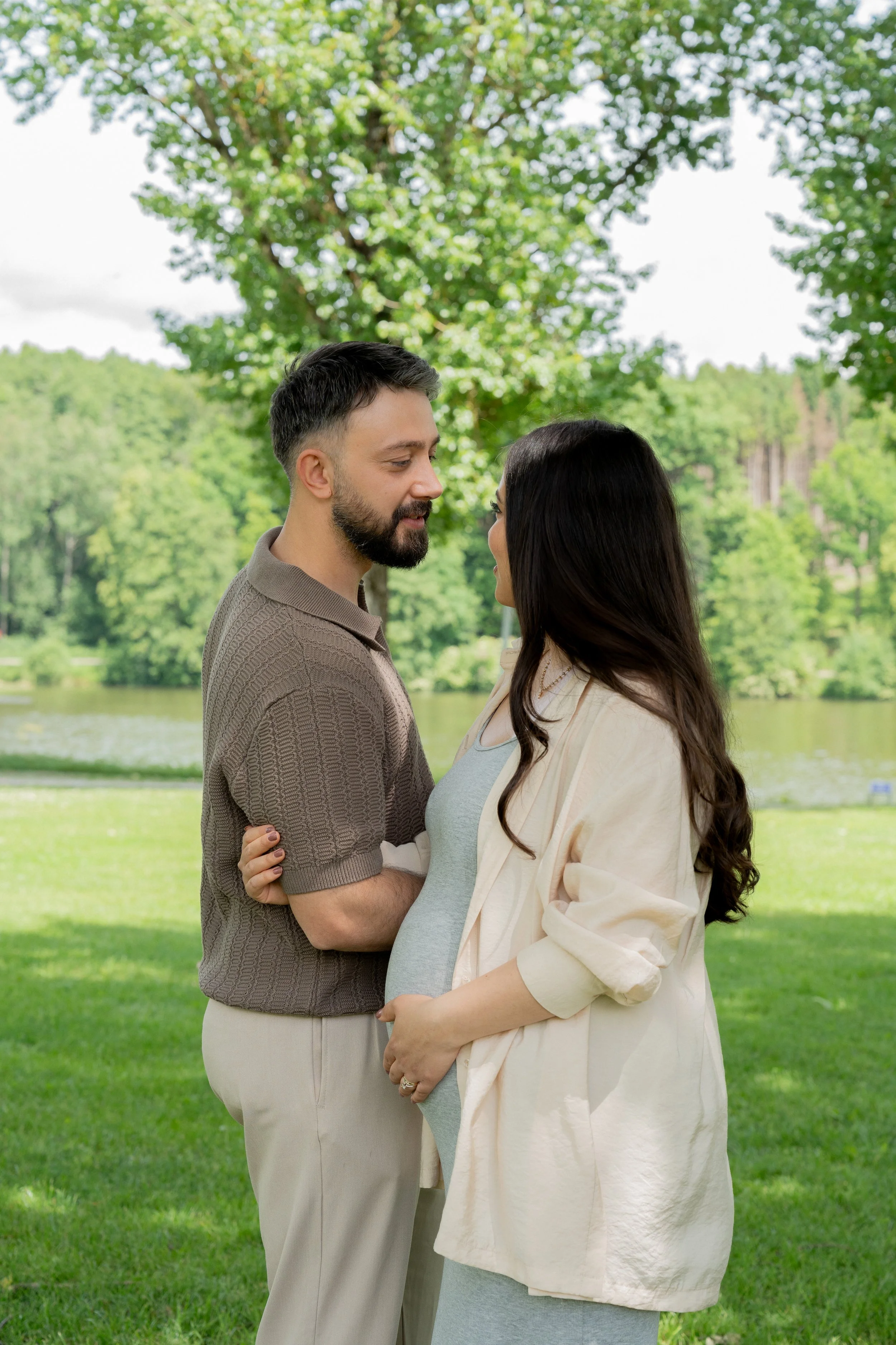 Ein schwangeres Paar steht im Park, schaut sich liebevoll an, im Hintergrund Bäume, Wasser und grünes Gras.