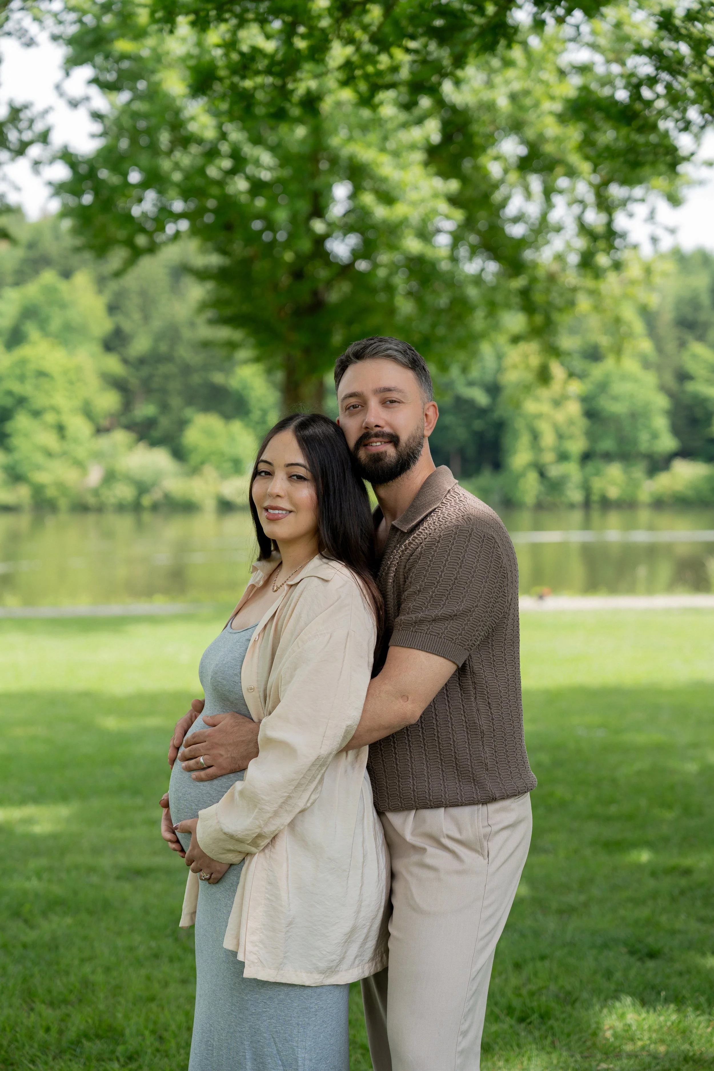 Ein Schwangertes Paar steht neben einem Baum in einem Park am See.