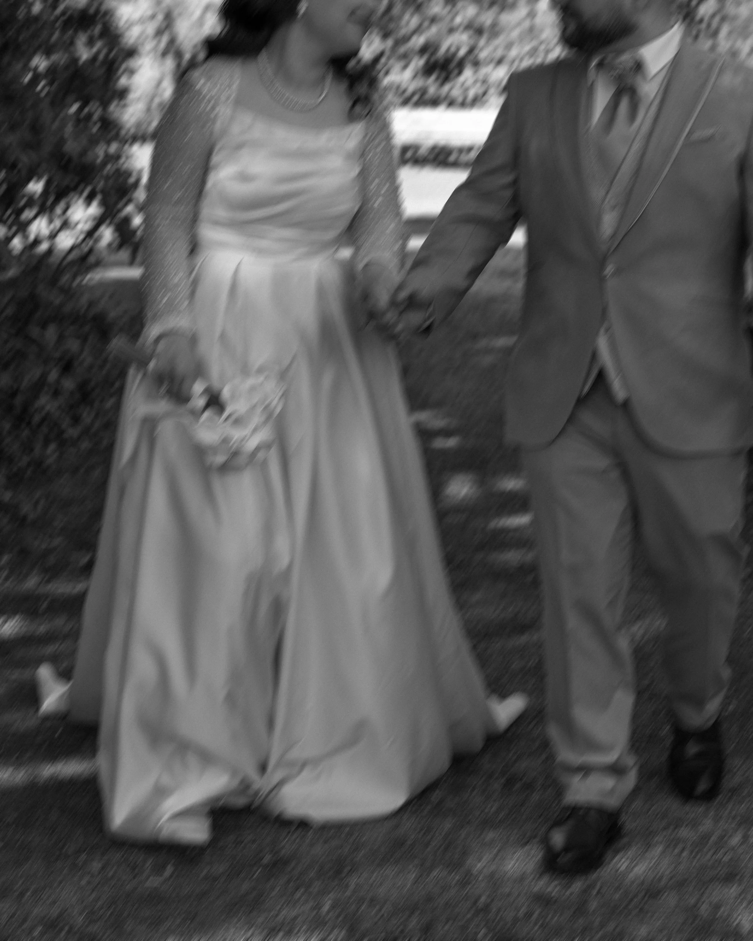 Ein Brautpaar geht Hand in Hand. Die Braut trägt ein langes, weißes Kleid und hält einen Blumenstrauß. Der Bräutigam ist in einem Anzug gekleidet. Schwarz-Weiß-Foto.