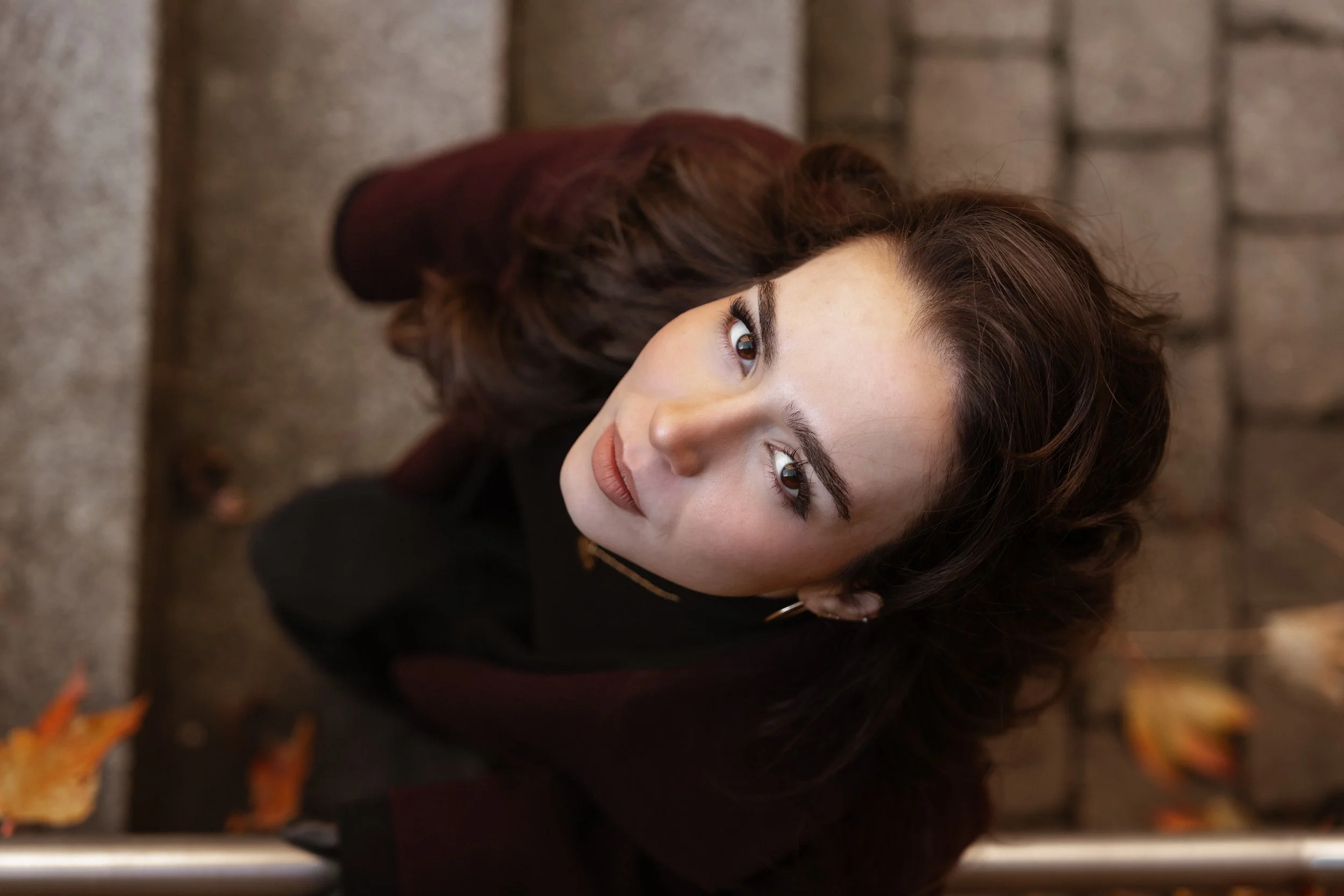 Nahaufnahme eines jungen Mädchens mit braunen, lockigen Haaren, die nach oben schaut, auf einem Kopfsteinpflaster mit herabgefallenen Herbstblättern.