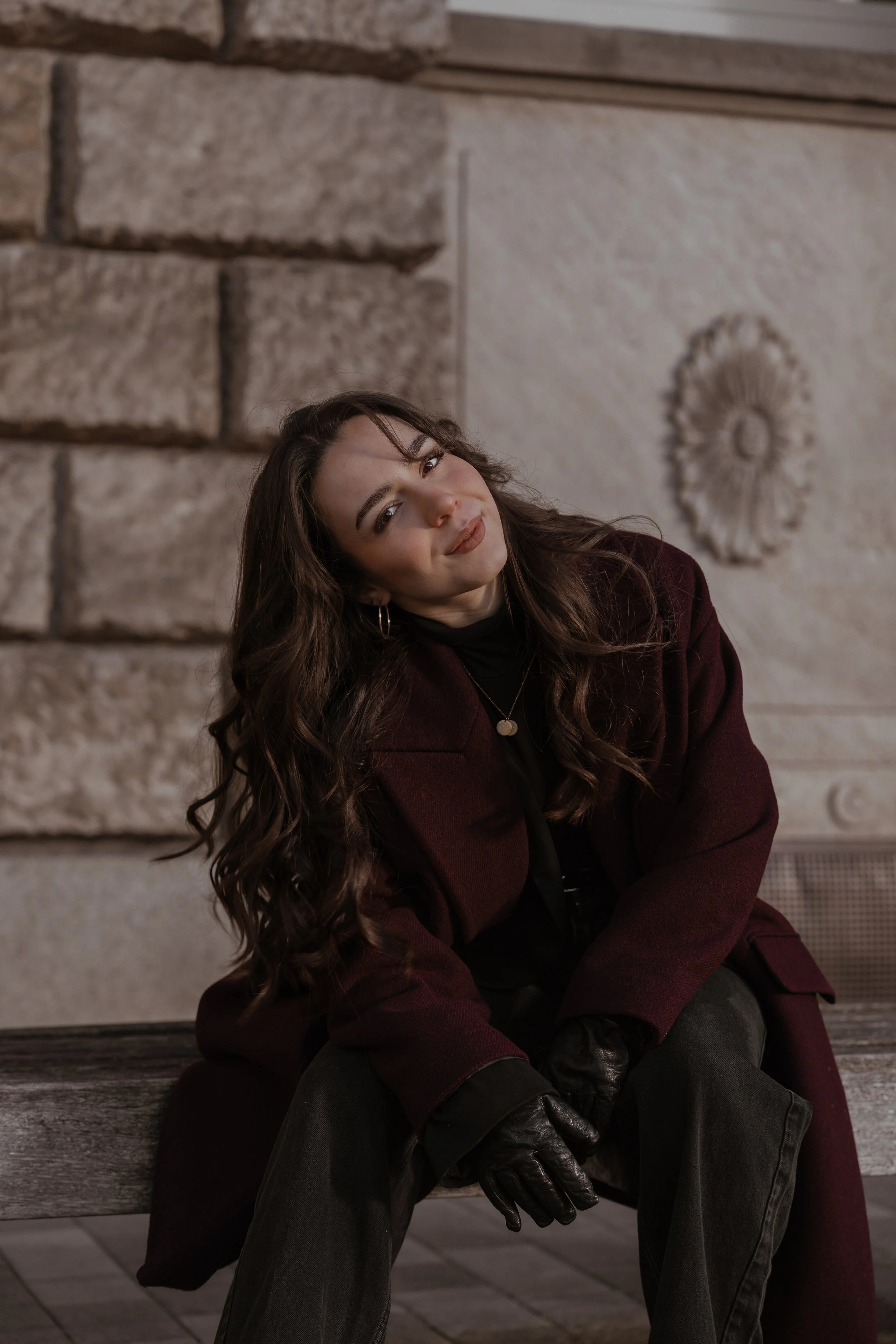 Junge Frau mit longem braunem Haar, die auf einer Holzbänke sitzt und lächelt. Sie trägt eine dunkle Jacke, schwarzen Pullover, schwarze Lederhandschuhe und Schmuck, vor einem Steinmauer mit dekorativen Elementen.