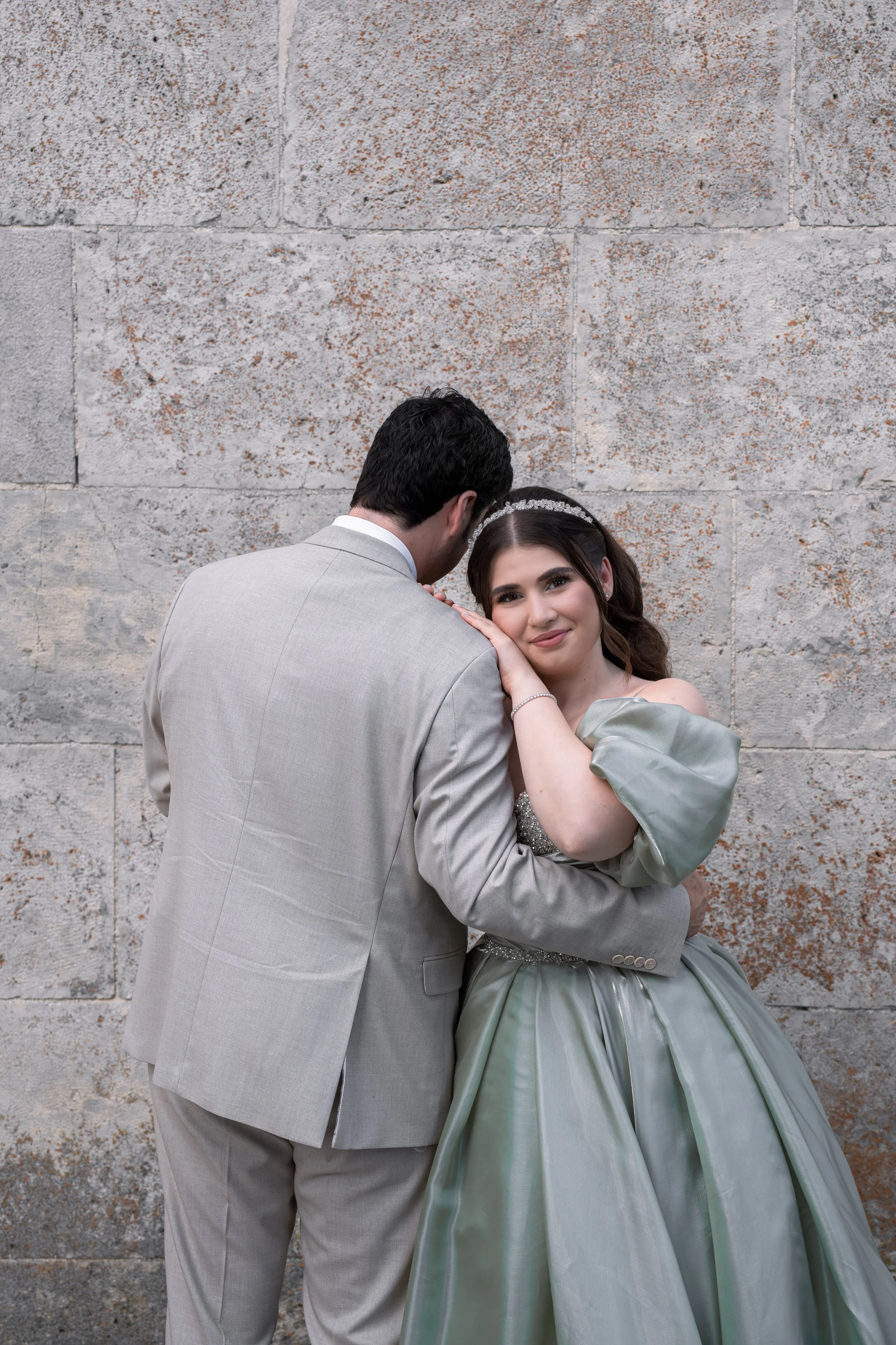 Ein Brautpaar steht vor einer Steinmauer, das Mädchen trägt ein langes, hellgraues Brautkleid mit Puffärmeln und Bandeau-Halsschleife, der Mann trägt einen hellgrauen Anzug.