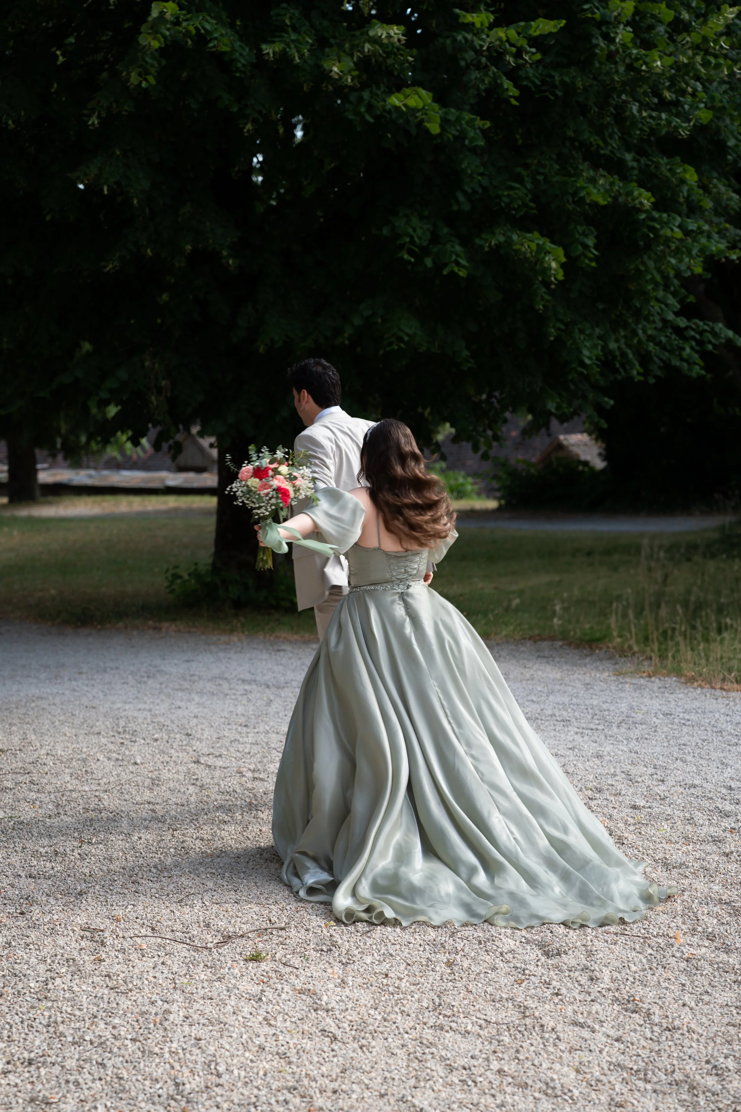 Ein Brautpaar steht im Freien, die Braut trägt ein ellenlanges, seidiges Hochzeitskleid und hält einen Blumenstrauß, während der Bräutigam in einem weißen Hemd und Anzugjacke steht.