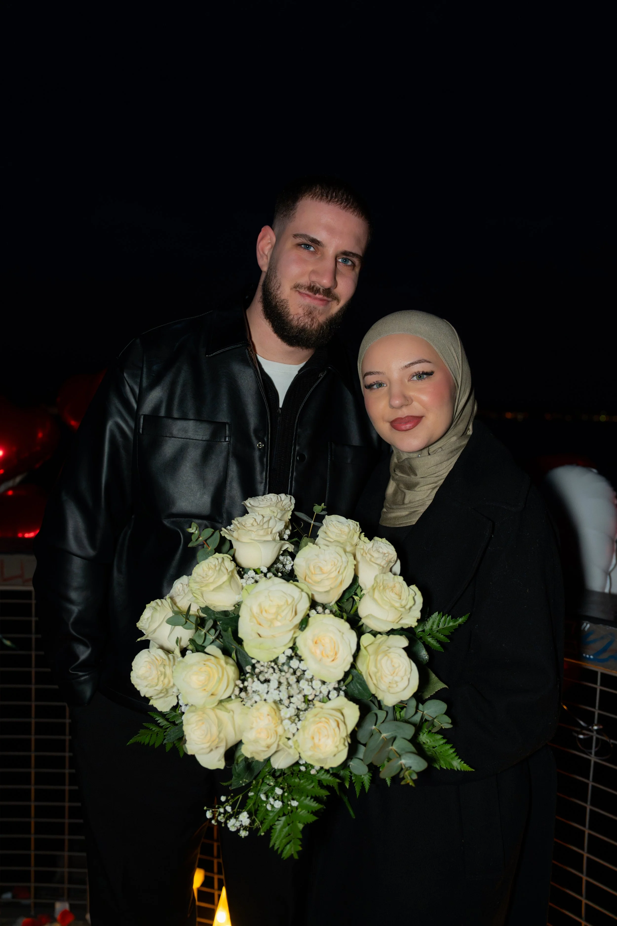Ein glückliches Paar auf einem Dach bei Nacht, das einen Blumenstrauß aus weißen Rosen hält.