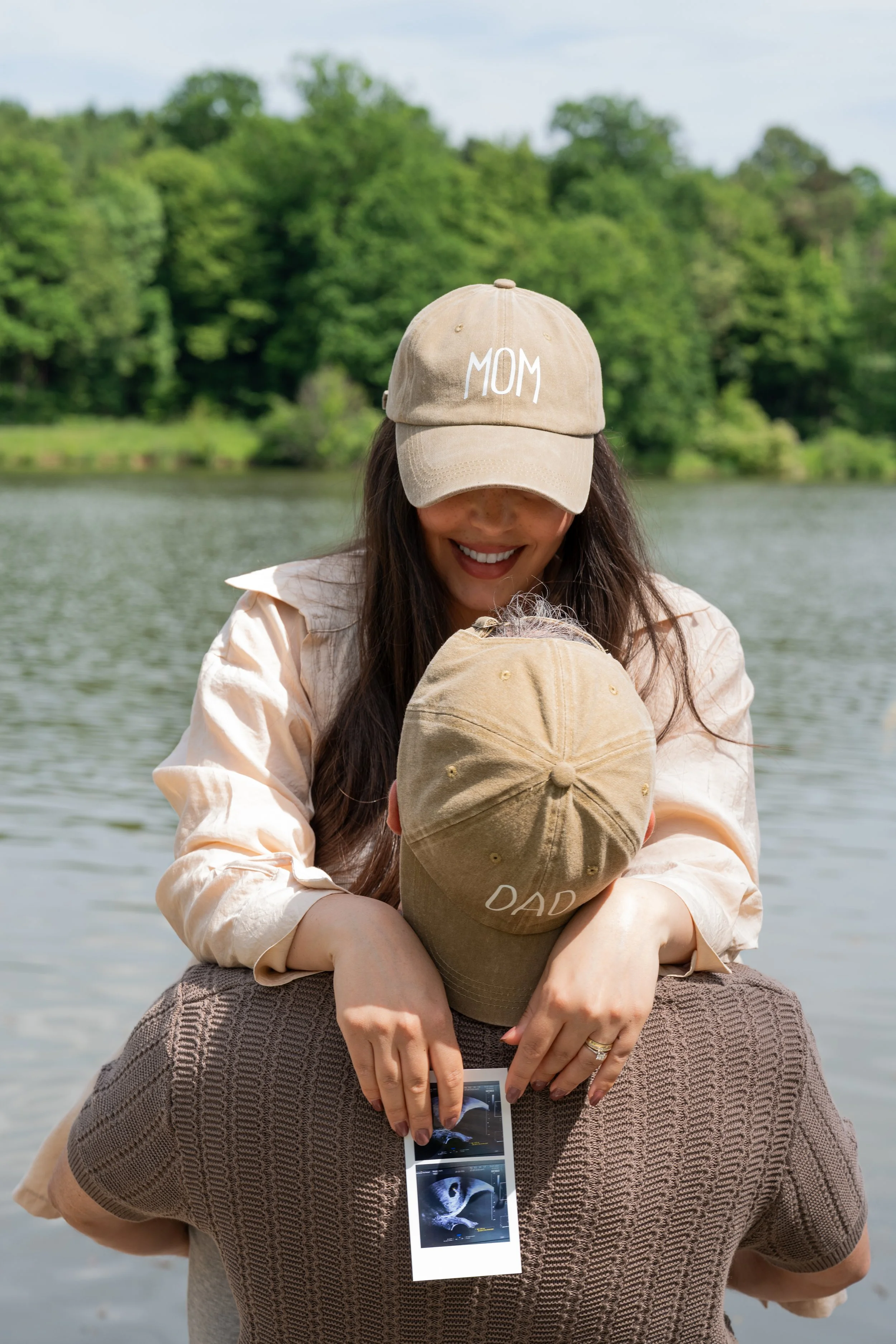 Frau schenkt Mann eine Umarmung am See, beide tragen beige Caps mit Beschriftung, sie ist fröhlich, er trägt Ultraschallbilder in der Hand.