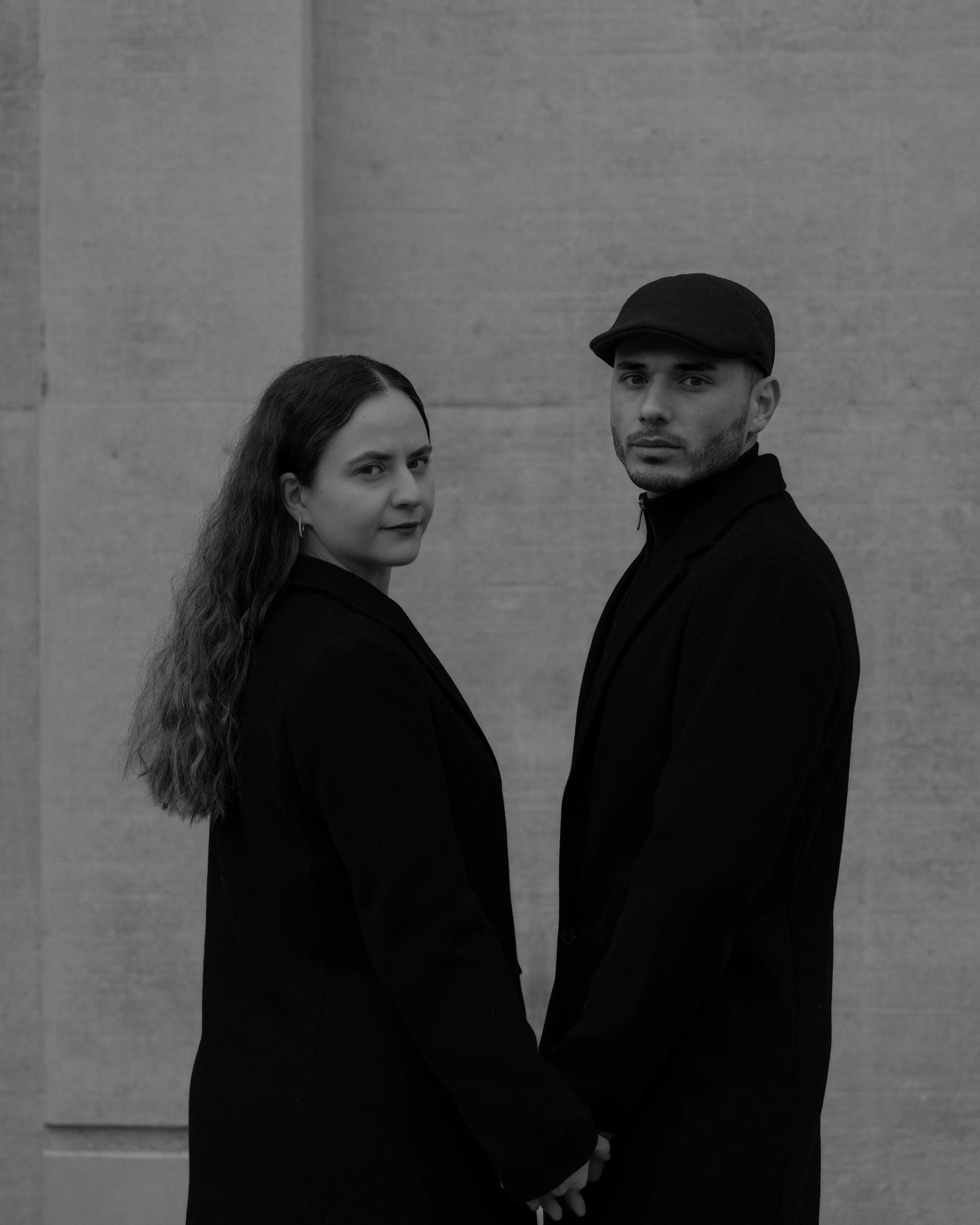 Zwei Personen in dunkler Kleidung stehen voreinander und halten sich an den Händen, Blick zur Kamera. Hintergrund aus Beton.