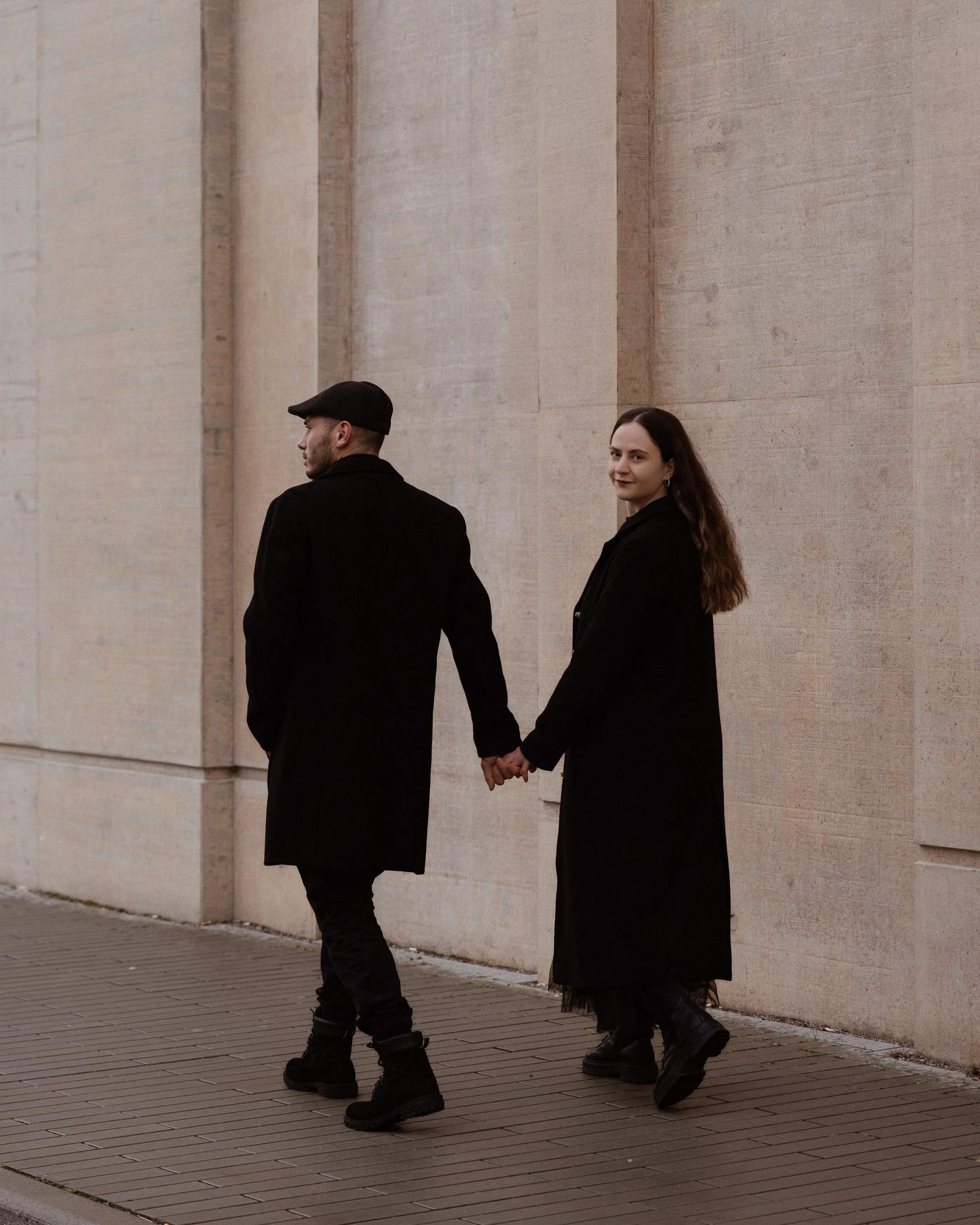 Ein Mann und eine Frau halten Händchen und gehen an einer Steinmauer entlang, beide tragen lange schwarze Mäntel und schwarze Schuhe, die Frau hat lange braune Haare.