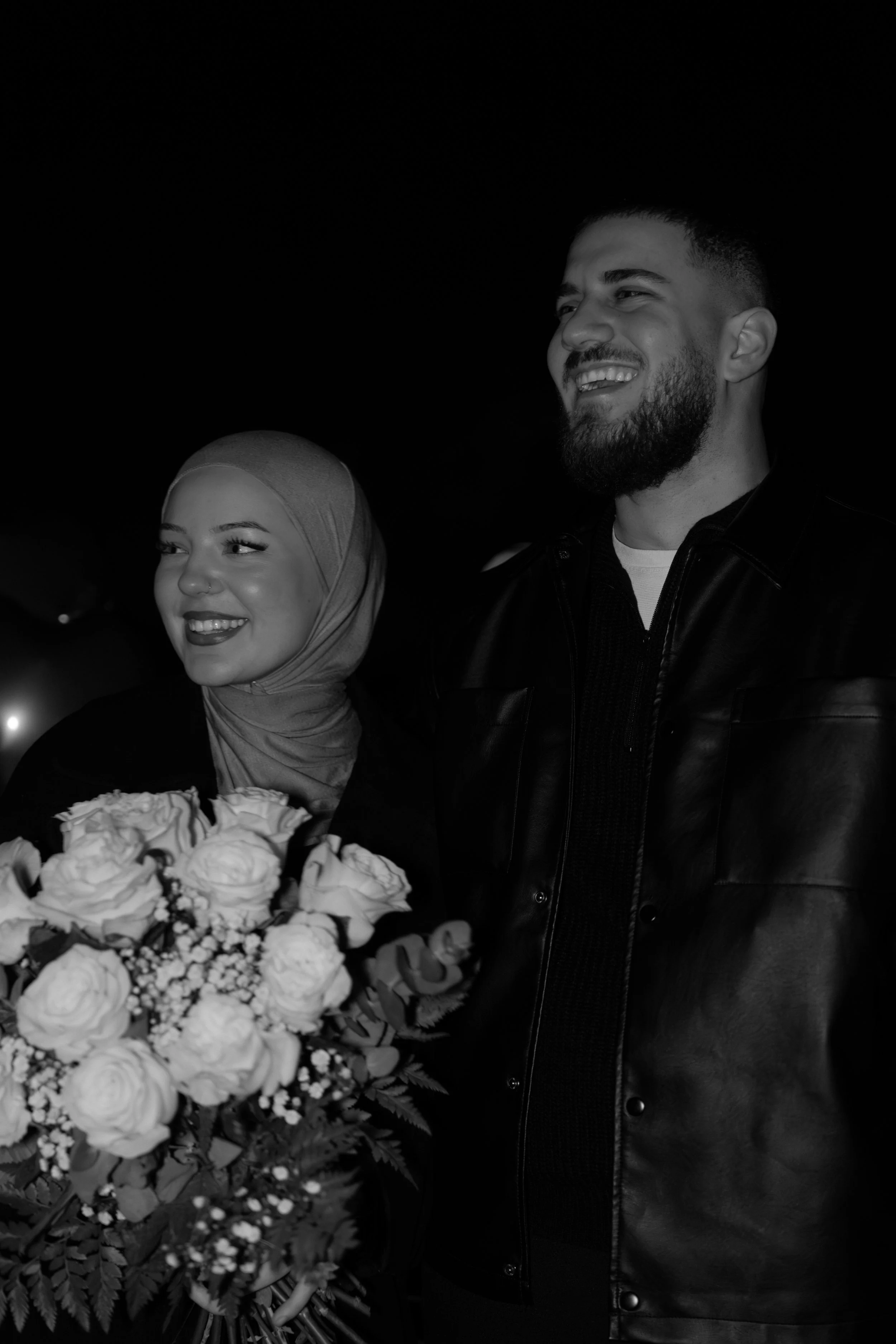 Ein lächelndes Paar steht in Nahaufnahme. Die Frau trägt ein Kopftuch und hält einen Blumenstrauß, der Mann hat einen Bart.