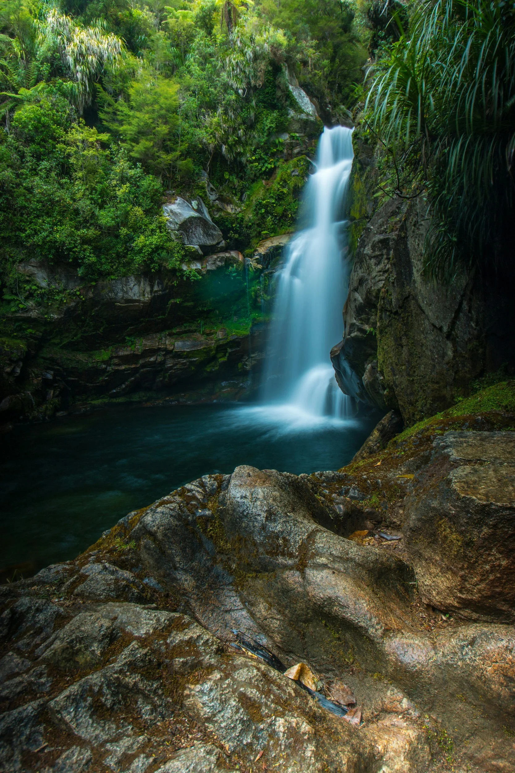 A multi-tiered waterfall cascading down rocks in a lush green forest, with stone steps on the right side for hiking.