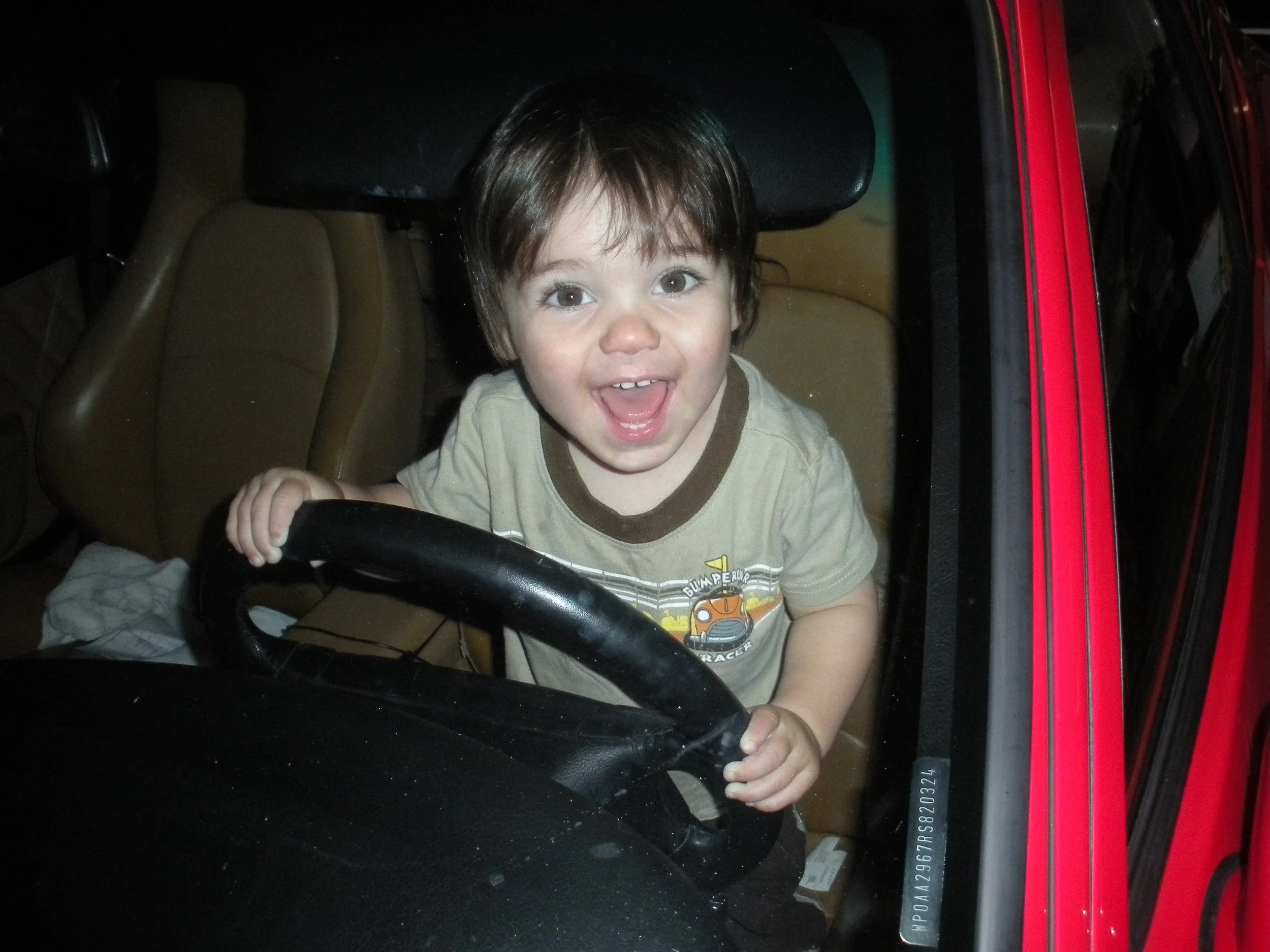 Smiling young boy with brown hair holding a car steering wheel inside a vehicle