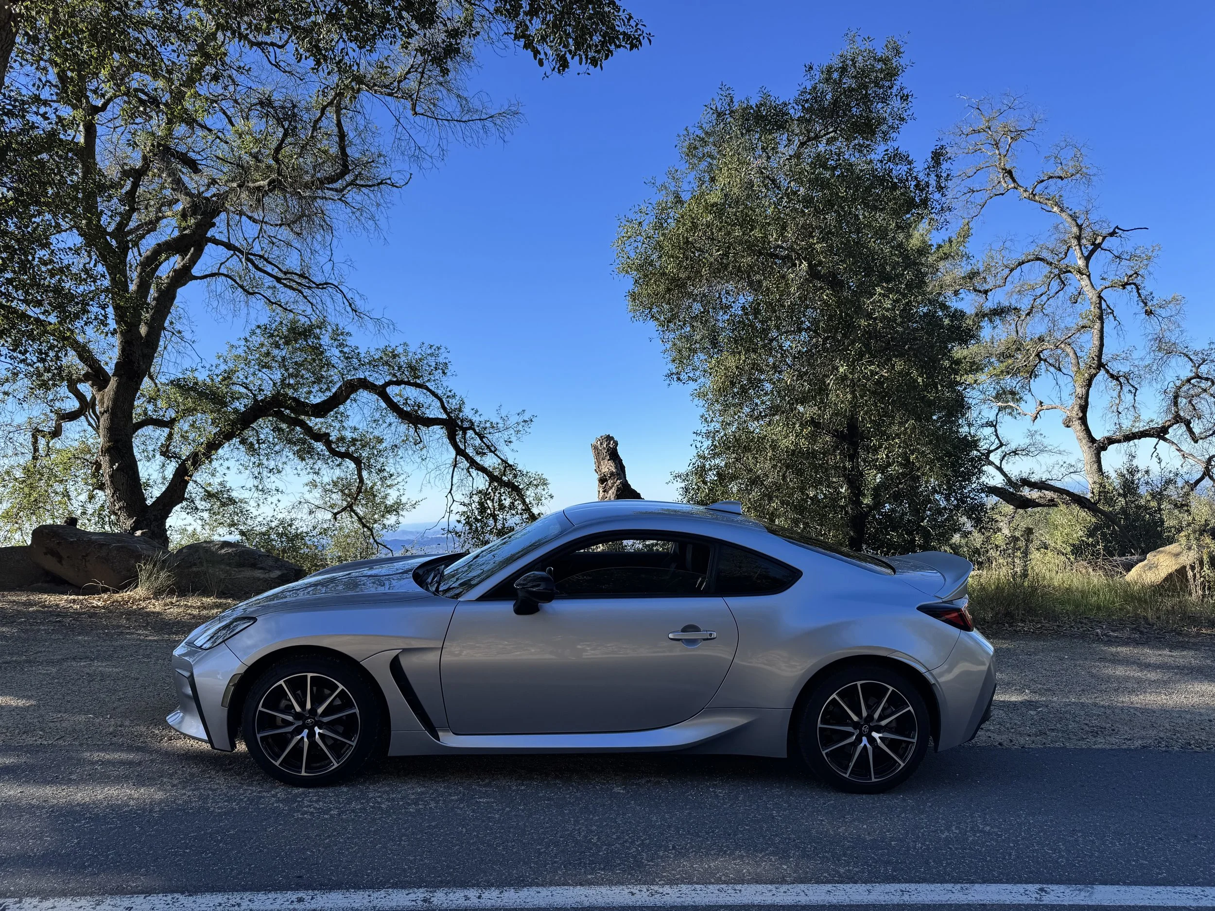 A silver sports car parked on a dirt road with trees and blue sky in the background.