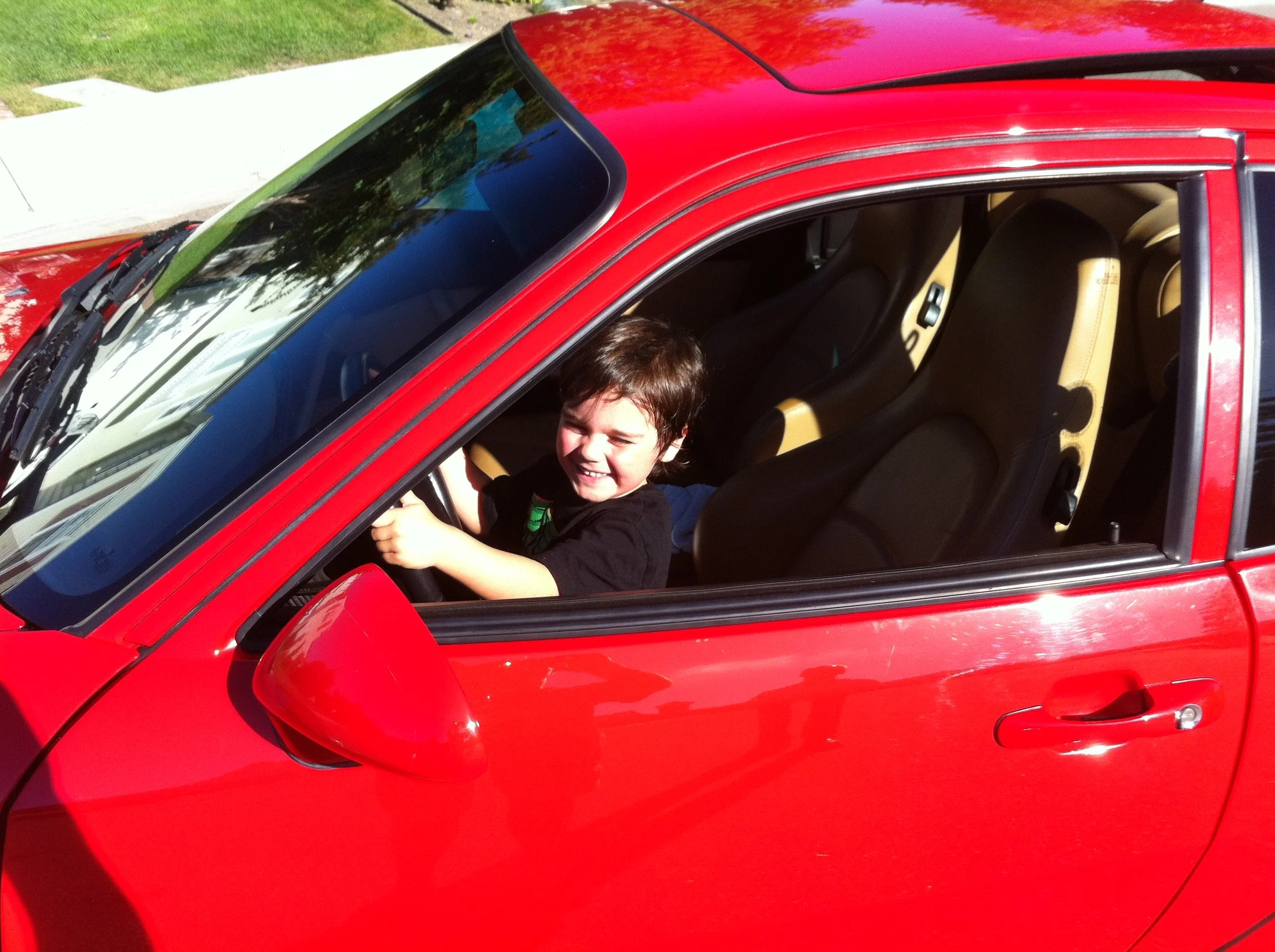 A young boy sitting in the driver's seat of a red car, smiling and holding the steering wheel.