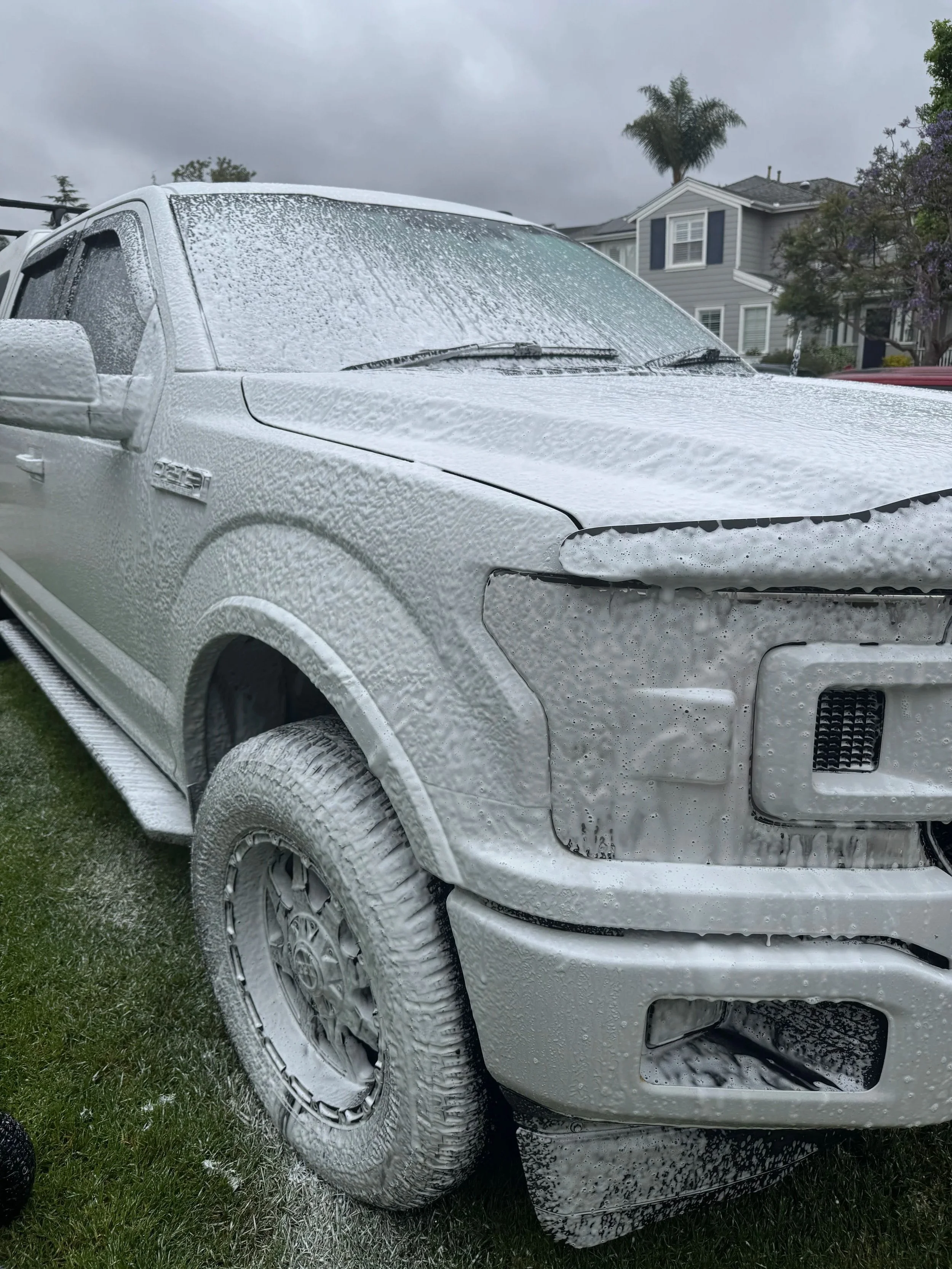 A silver pickup truck covered in white foam, likely from a car wash, parked on a grassy area with residential houses and trees in the background.
