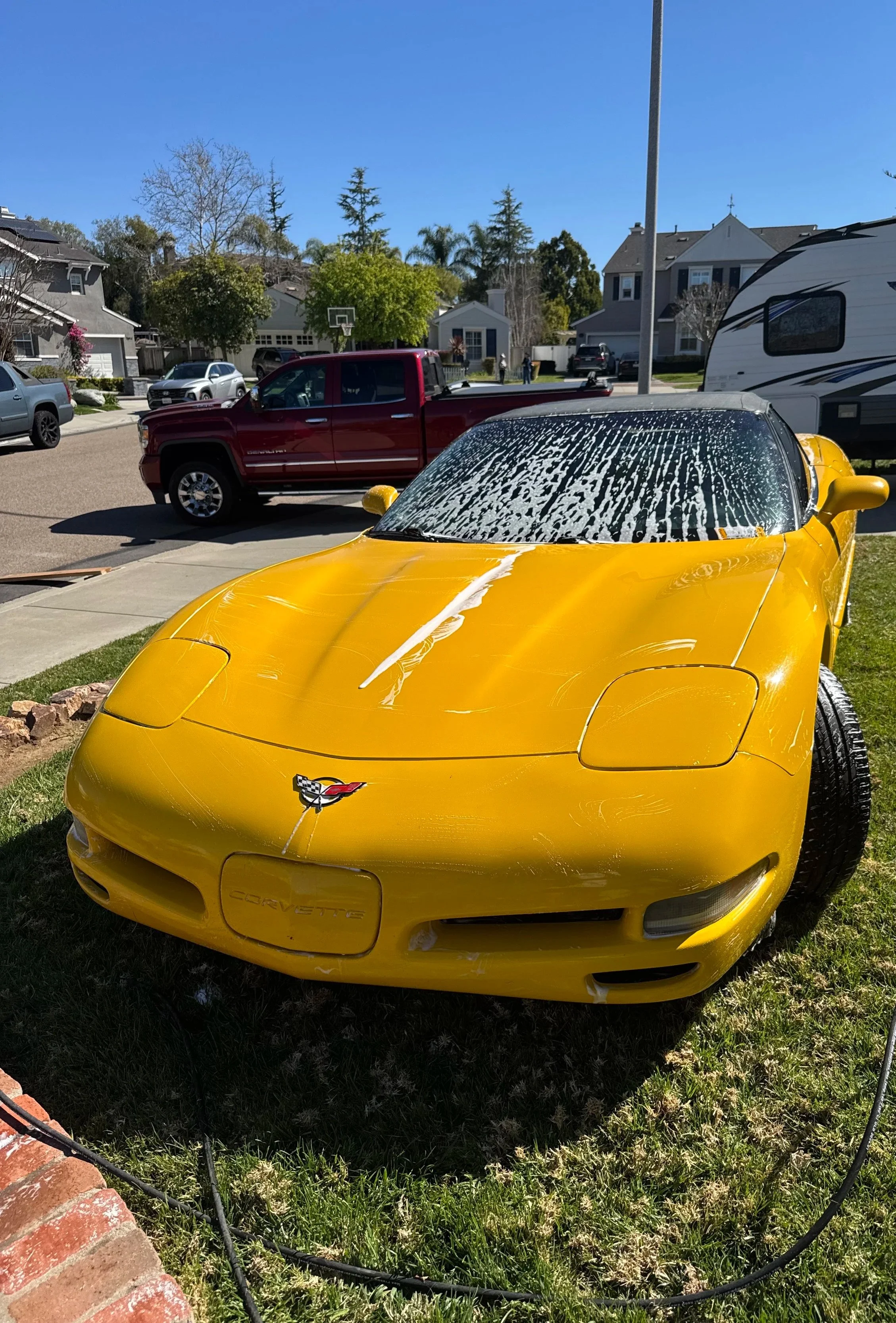 A yellow Chevrolet Corvette with foam on the windshield, parked on a grassy area in front of houses on a sunny day.